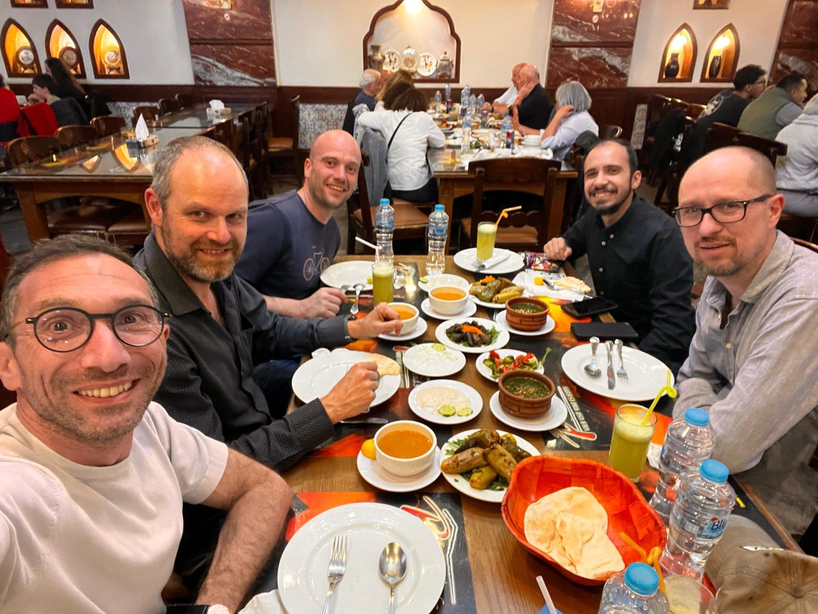 Group of six men sitting around a table in a restaurant, enjoying a meal with various dishes, drinks, and condiments. They are smiling and looking at the camera, with a background of other diners and restaurant decor.