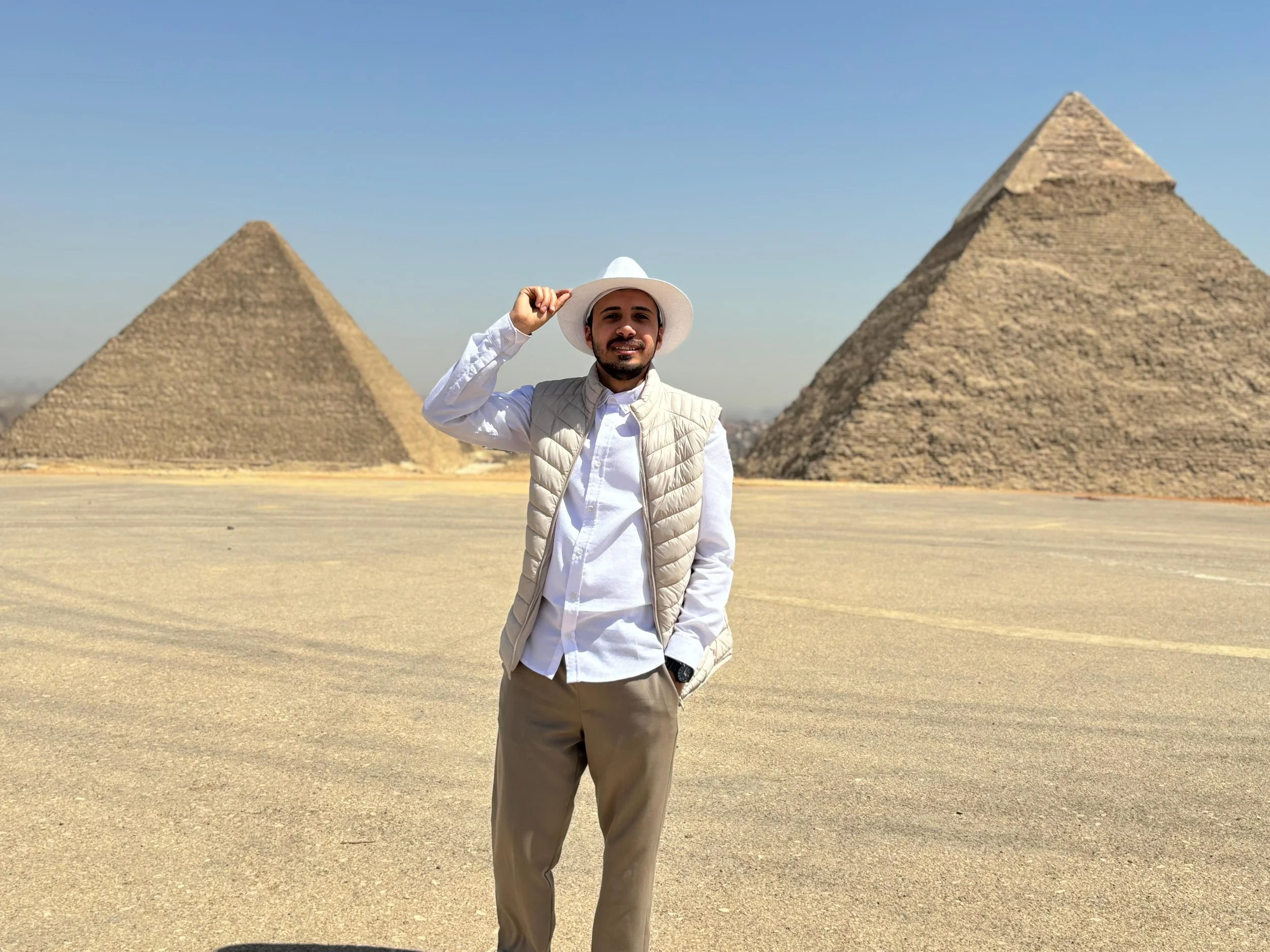 A man standing in front of the pyramid-shaped structures of the Egyptian pyramids, wearing a white fedora hat, white shirt, beige vest, and beige trousers, and smiling.