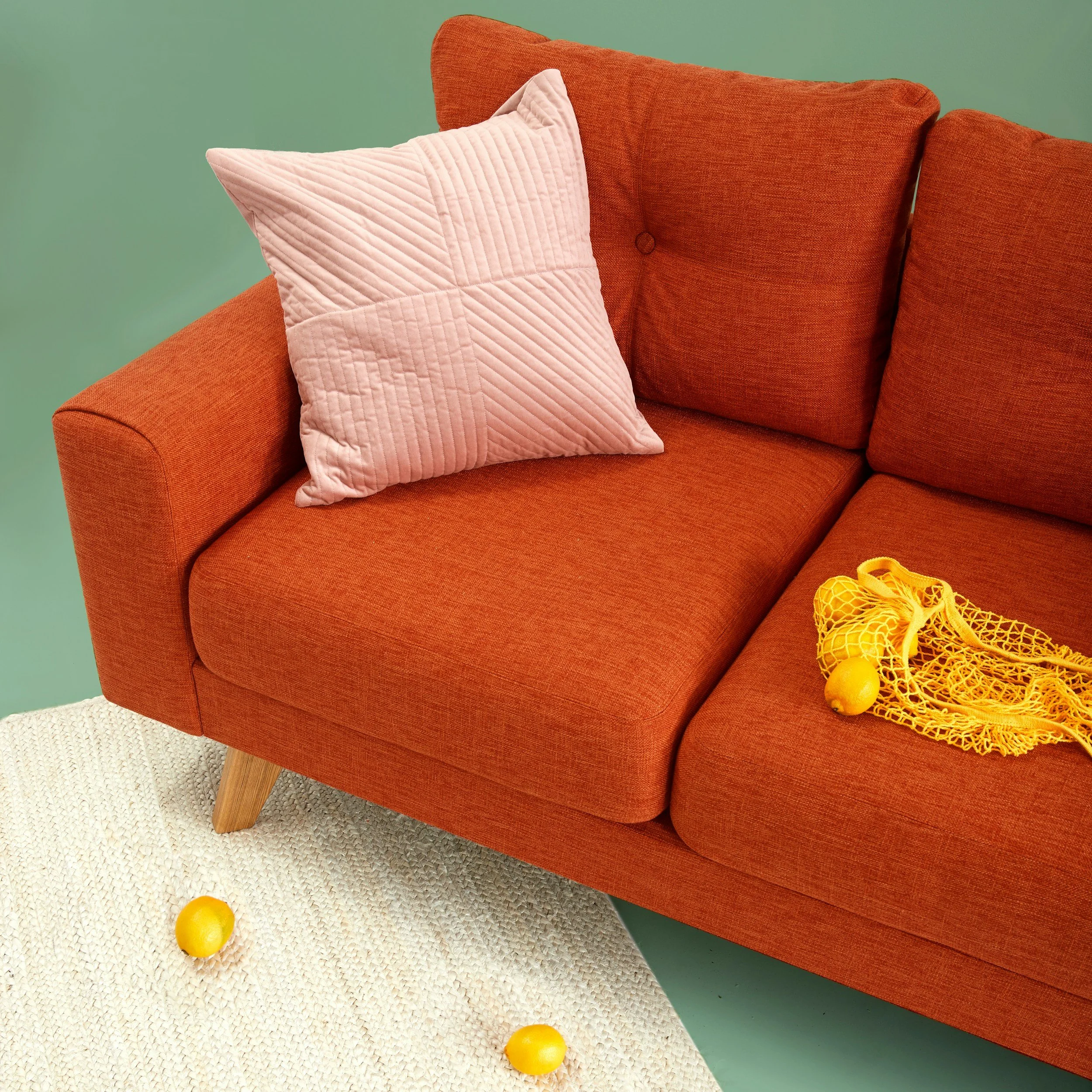 Close-up of an orange upholstered sofa with wooden legs, a pink quilted pillow on the left side, a green wall in the background, a ivory textured rug on the floor, and three yellow tennis balls, one on the rug and two on the sofa.