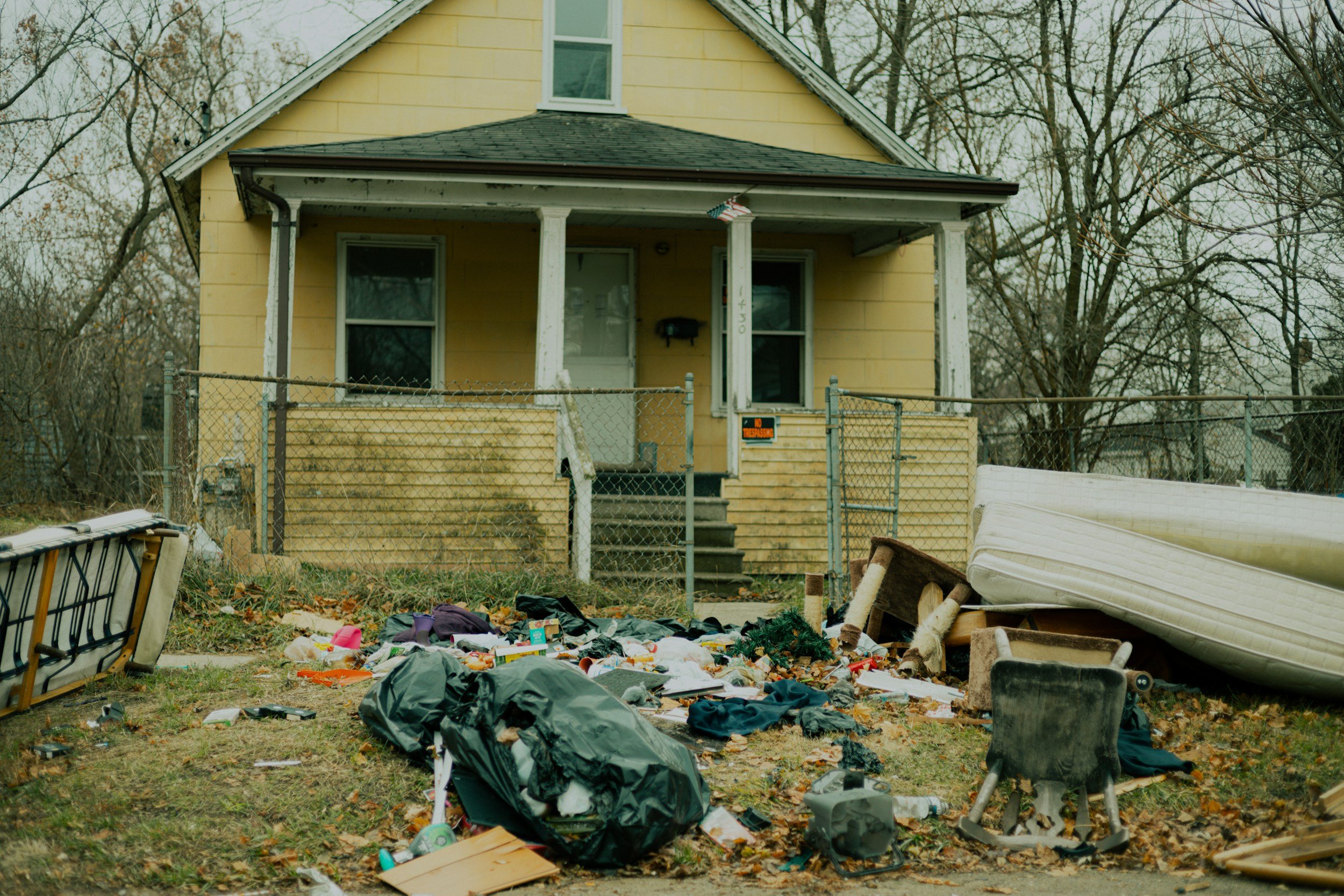 The front of a yellow house with a porch, surrounded by a chain-link fence, has clutter including trash bags, furniture, and scattered debris in the yard.