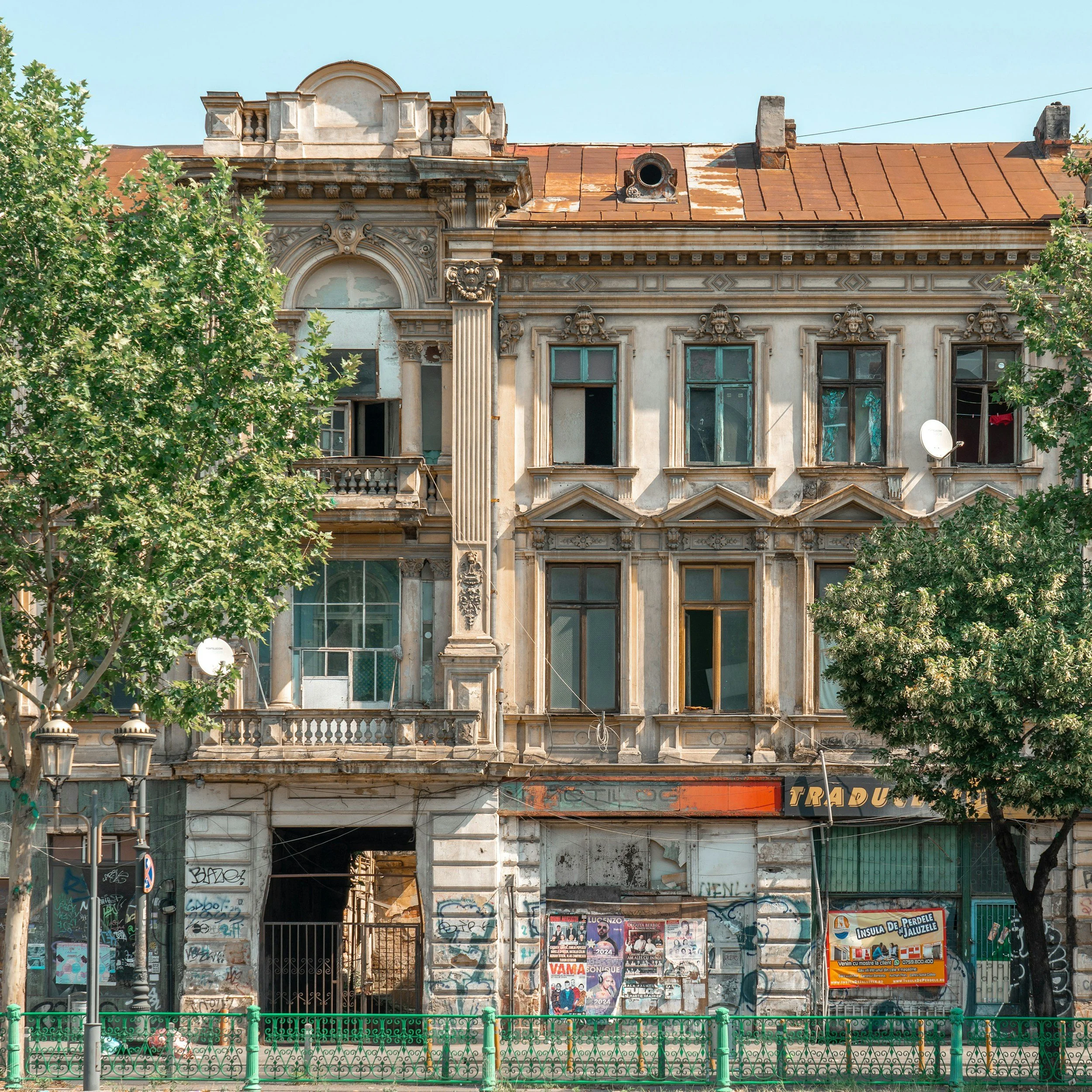 Old, abandoned building with classical architectural elements and graffiti on the lower part, partially obscured by two trees and a green fence in the foreground.