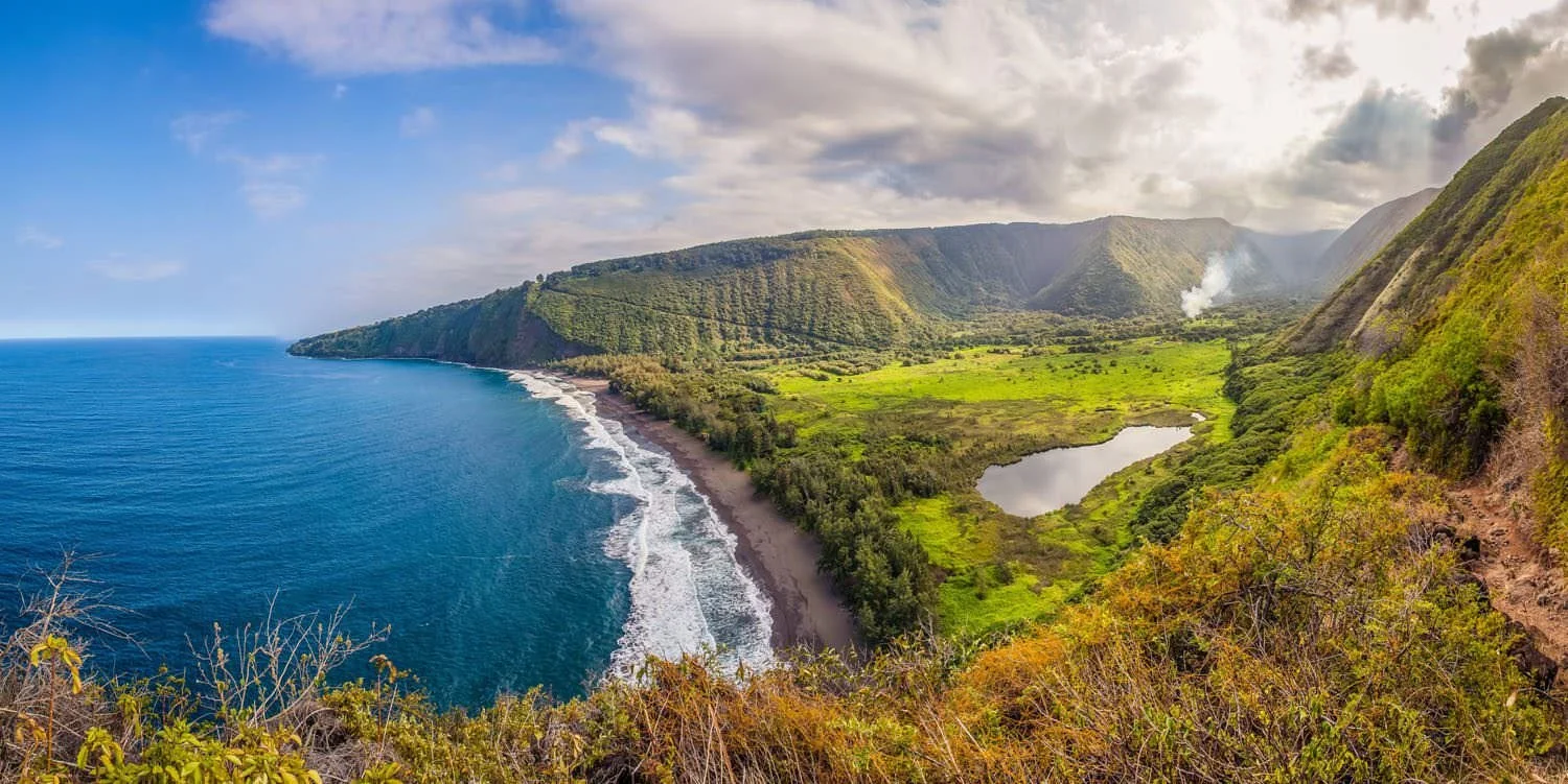 A coastal landscape with a sandy beach, ocean waves, green hills, and a cloudy sky.