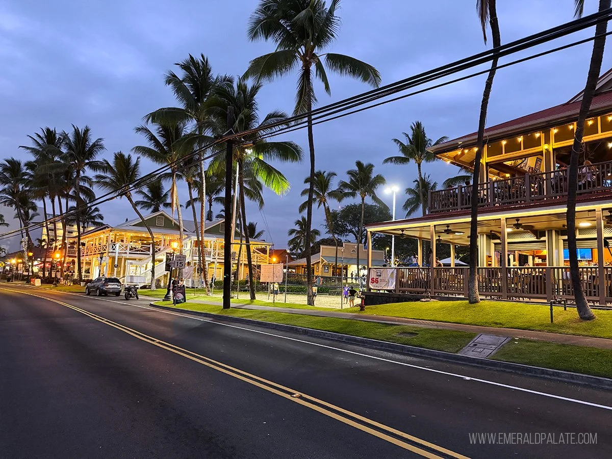A street scene at dusk with palm trees, illuminated restaurants, and a motorcycle on the road.