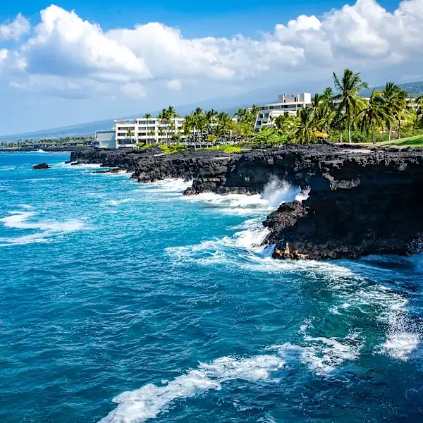 Oceanfront with waves crashing against black lava rocks, white modern resort buildings, palm trees, and cloudy sky in a tropical coastal area.