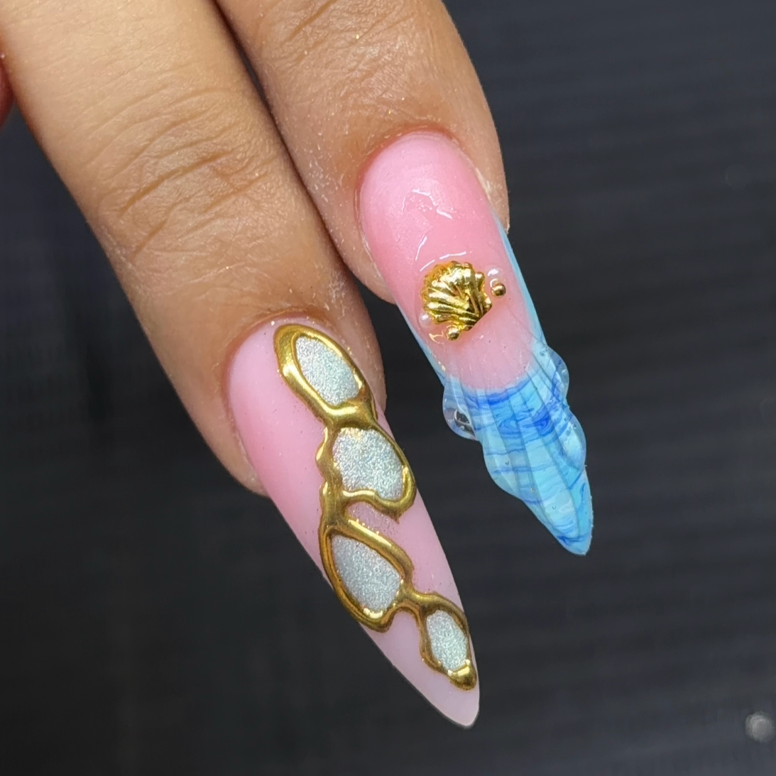 Nail art with pink base, gold seashell and pearl accents, and a blue and clear acrylic pointed extension resembling a seashell.