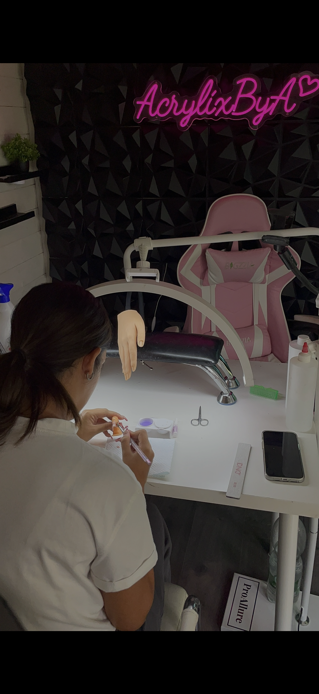 Woman performing a manicure at a nail salon workstation with nail tools and a pink massage chair in the background, illuminated by a neon pink sign reading 'AcrylixByA'.