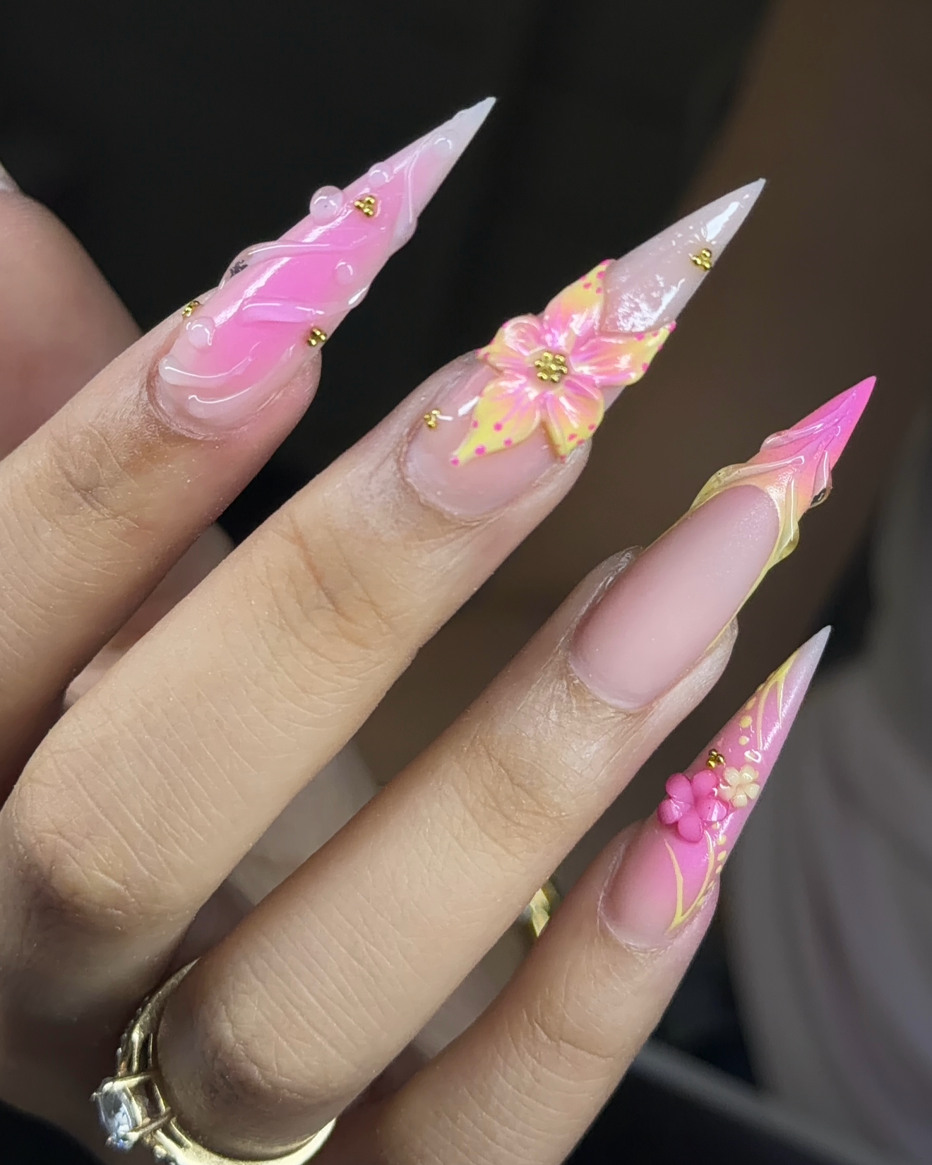 Close-up of a hand with long, decorated acrylic nails featuring pink, white, and yellow colors in floral and swirl designs, adorned with small gold beads and tiny 3D flower accents.