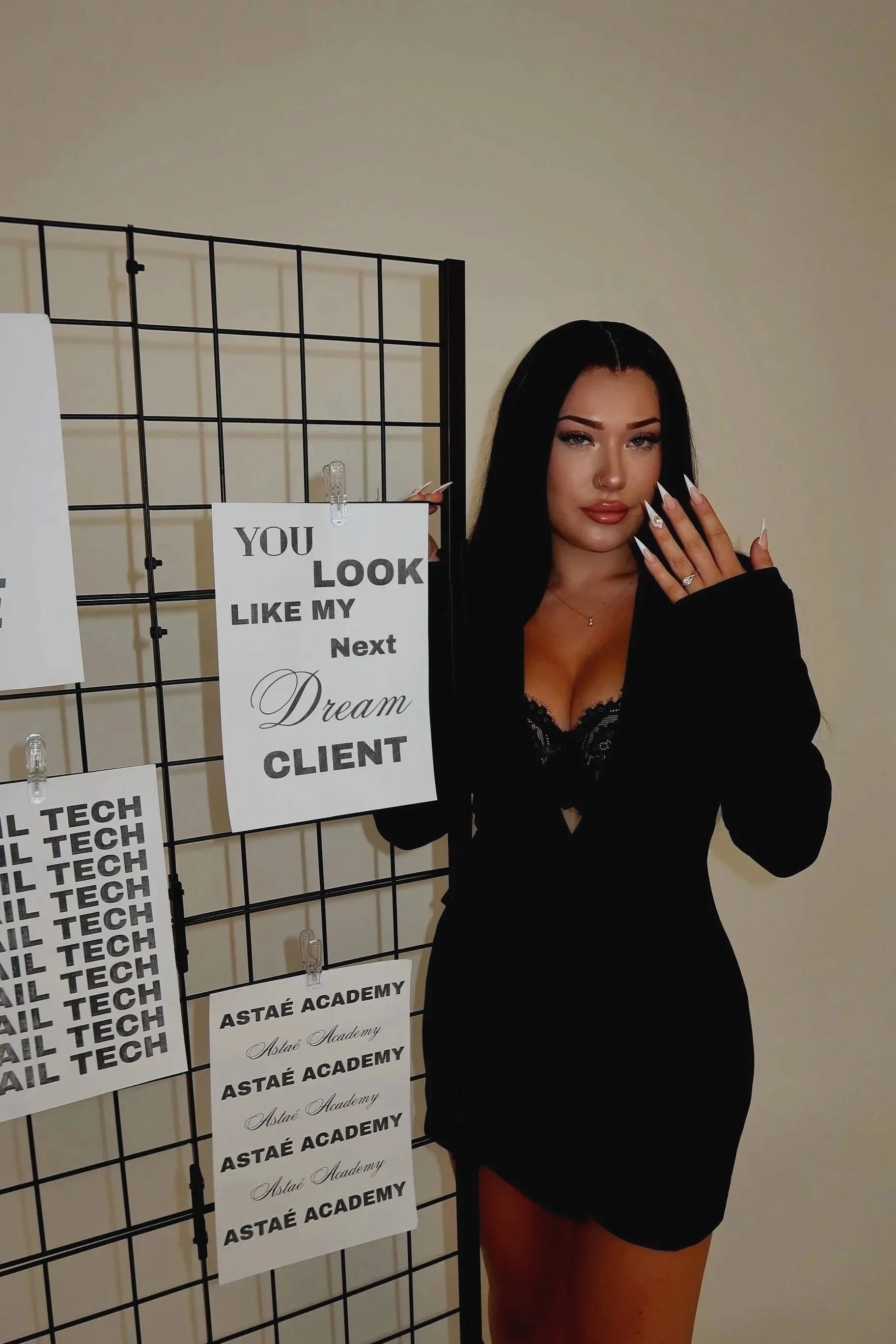 A woman with long black hair, makeup, and a nose piercing posing next to a display of signs with various texts, including 'You look like my next dream client' 'Academy.' She is wearing a black lace top and matching black skirt, holding her left hand near her face showing rings and long white nails.