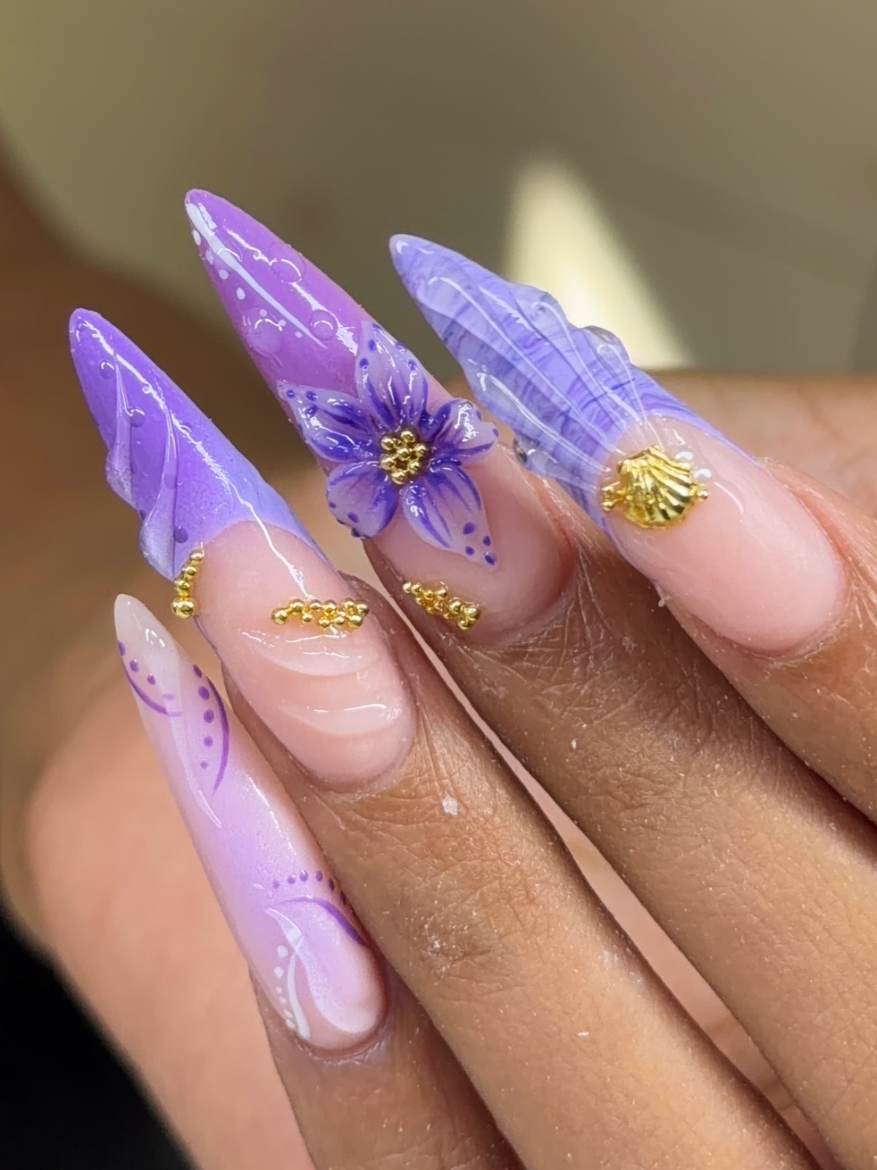 Close-up of a hand with elaborate purple nail art featuring painted flowers, gold embellishments, and 3D floral designs on long, pointed acrylic nails.