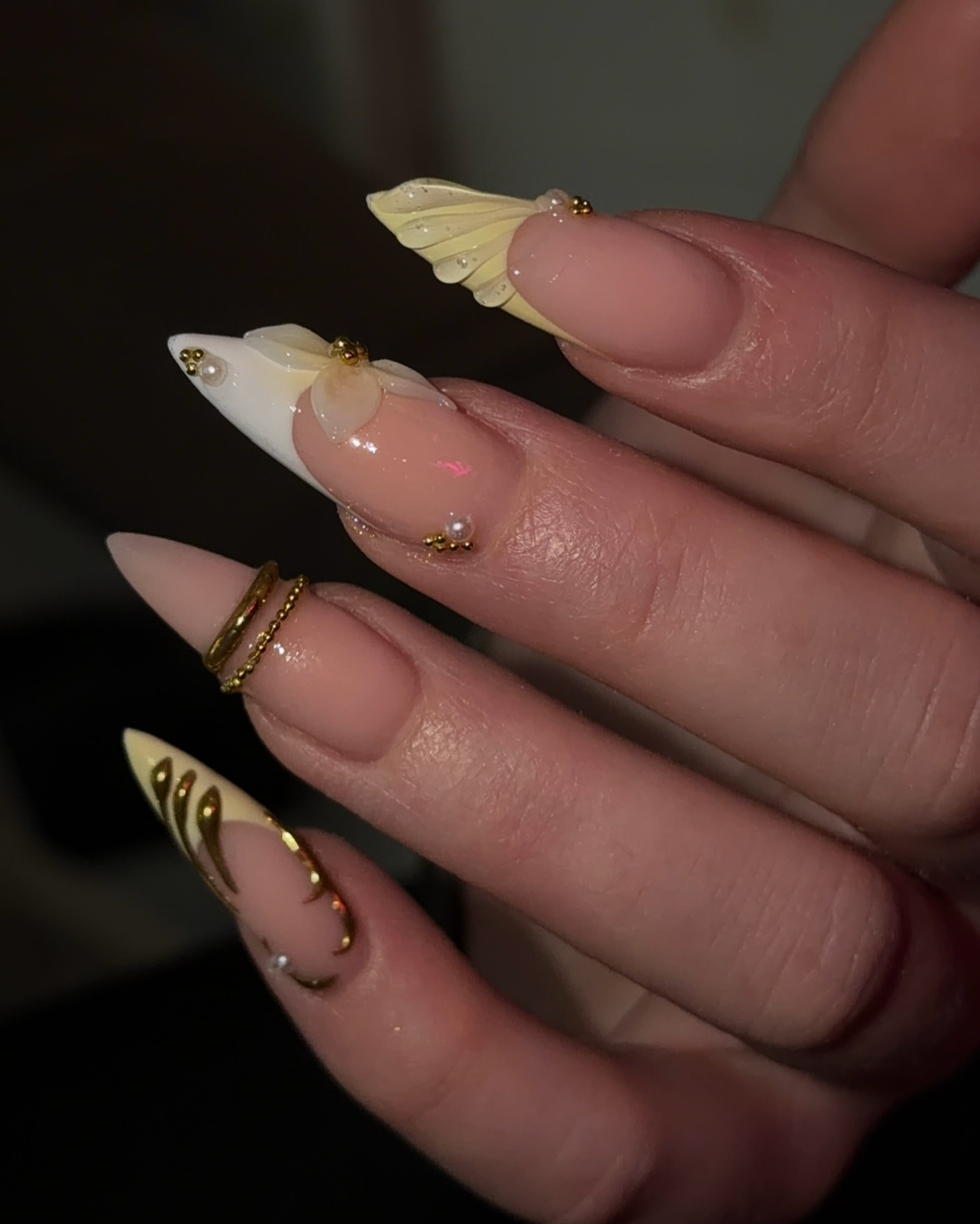 Close-up of a hand with decorated nails featuring 3D floral designs, pearls, and gold accents, with rings on the fingers.