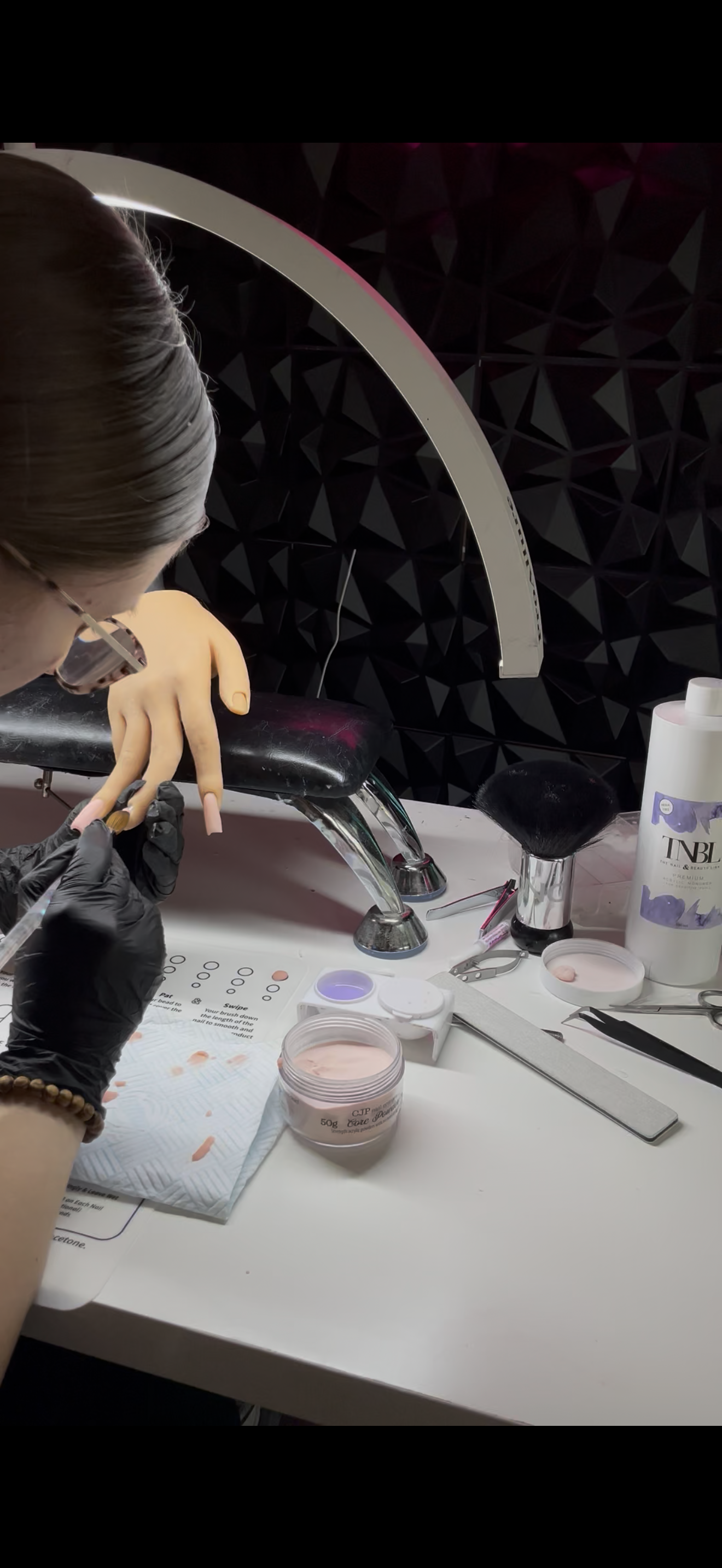 A person receiving a pink nail dip powder manicure at a salon station, with manicure tools, nail polish, and a lamp on the table.