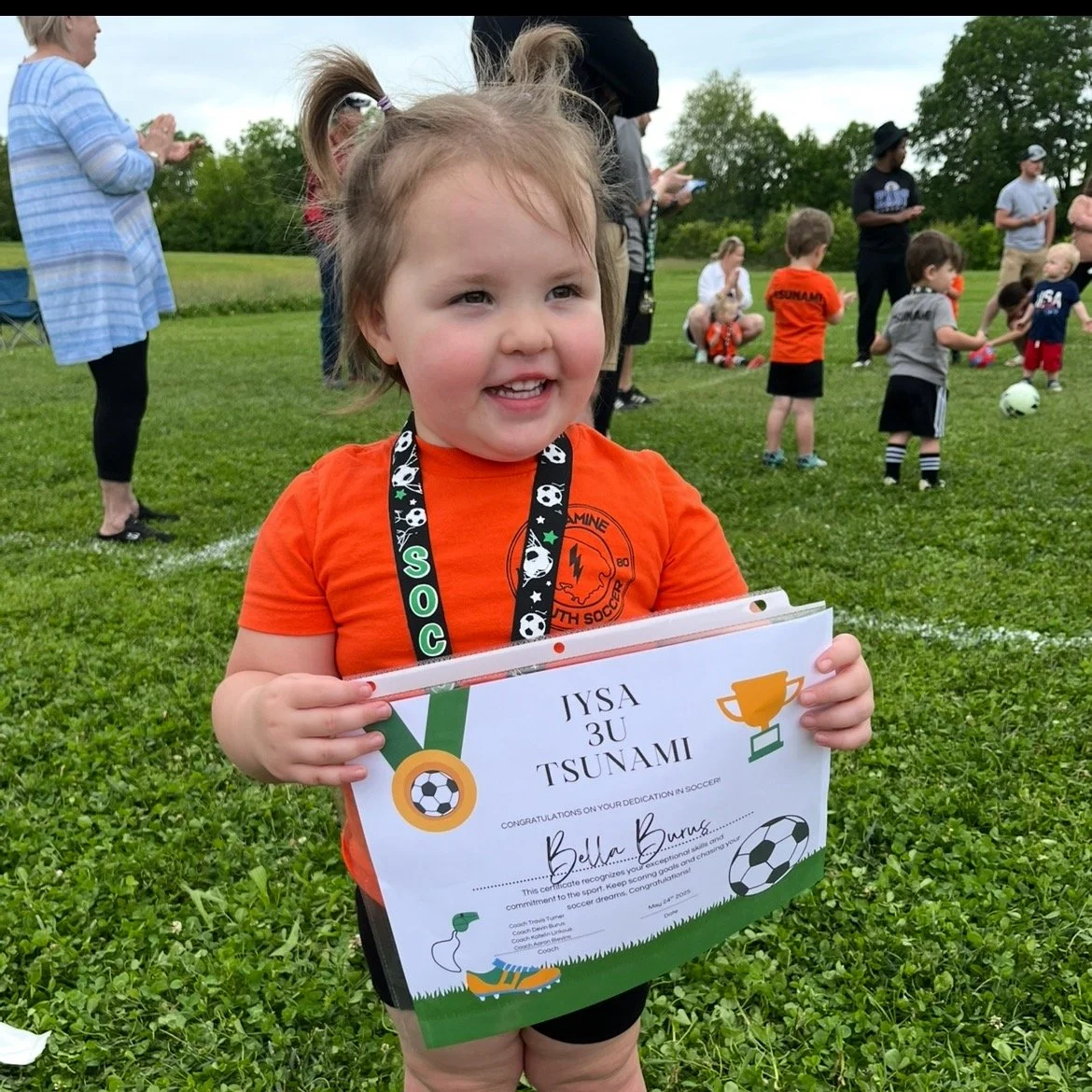 Bella Grace smiling proudly in her soccer uniform while holding a participation certificate, reflecting her joyful spirit and love for learning and new experiences.