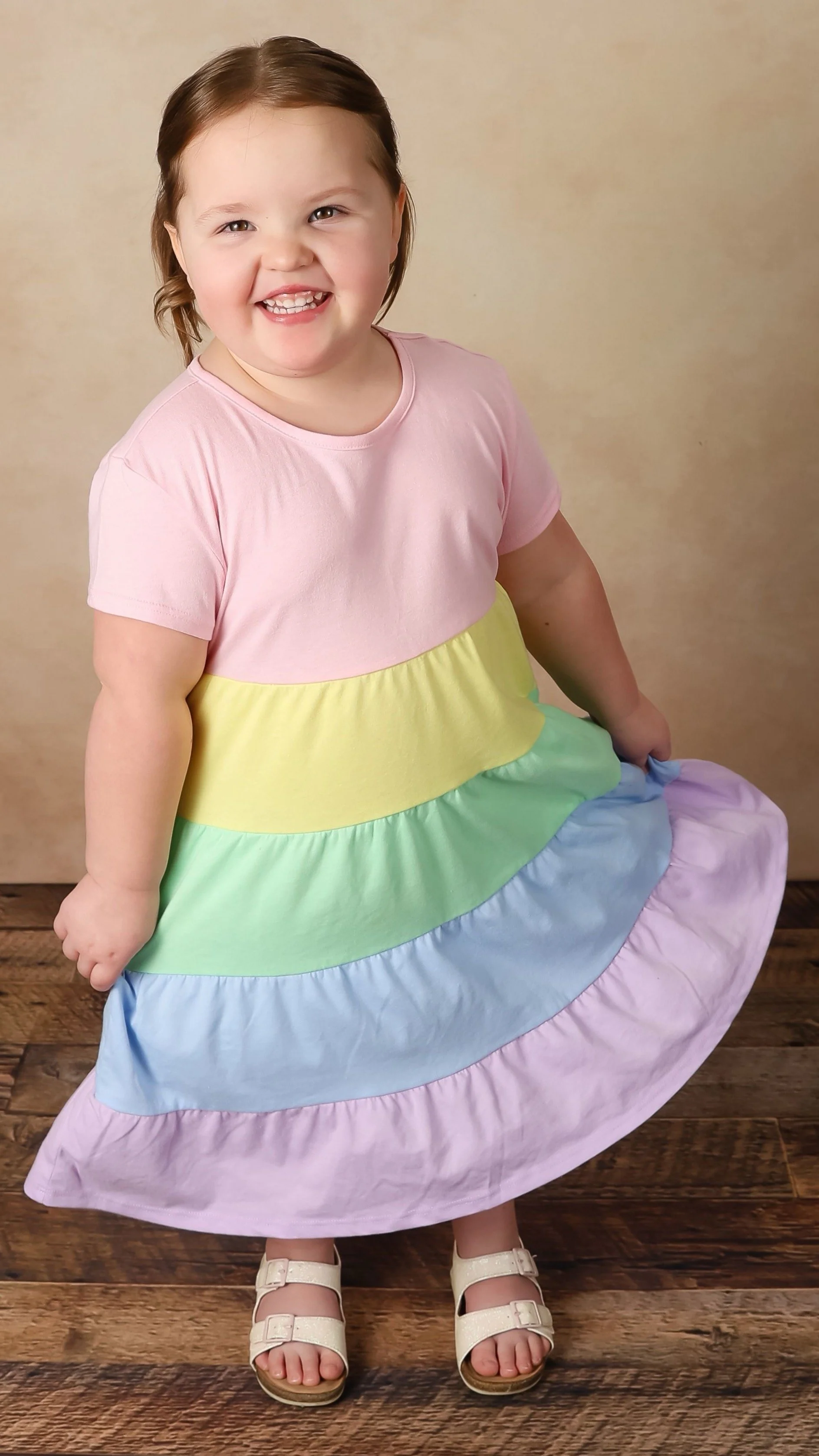 Bella Grace smiling joyfully in a rainbow-colored dress while standing outdoors. Her joyful expression reflects the childlike faith and hope that inspired the Believe Like Bella ministry.