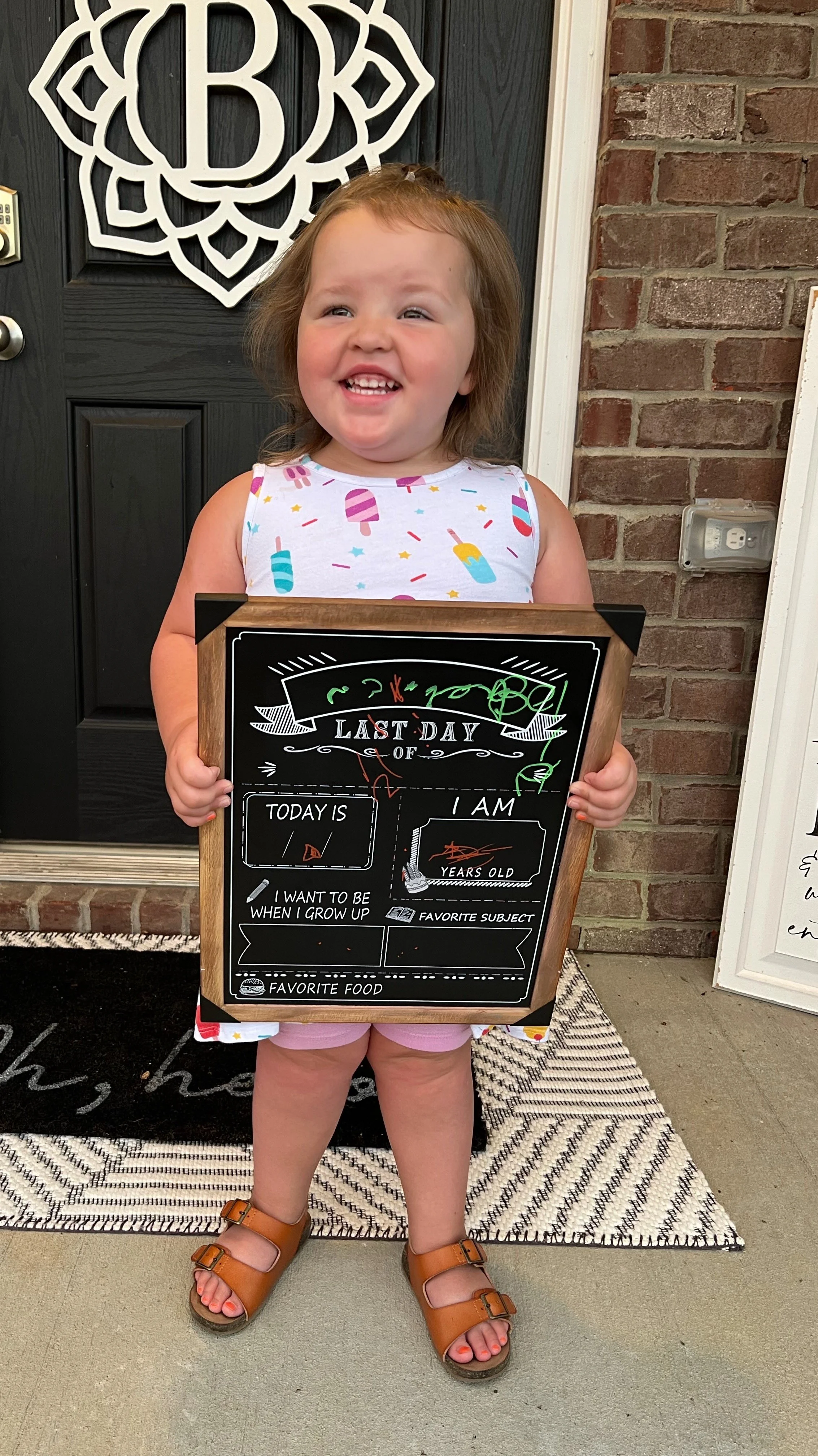 Bella Grace smiling on her first day of school while proudly holding her classroom sign, capturing her excitement, curiosity, and joyful spirit.