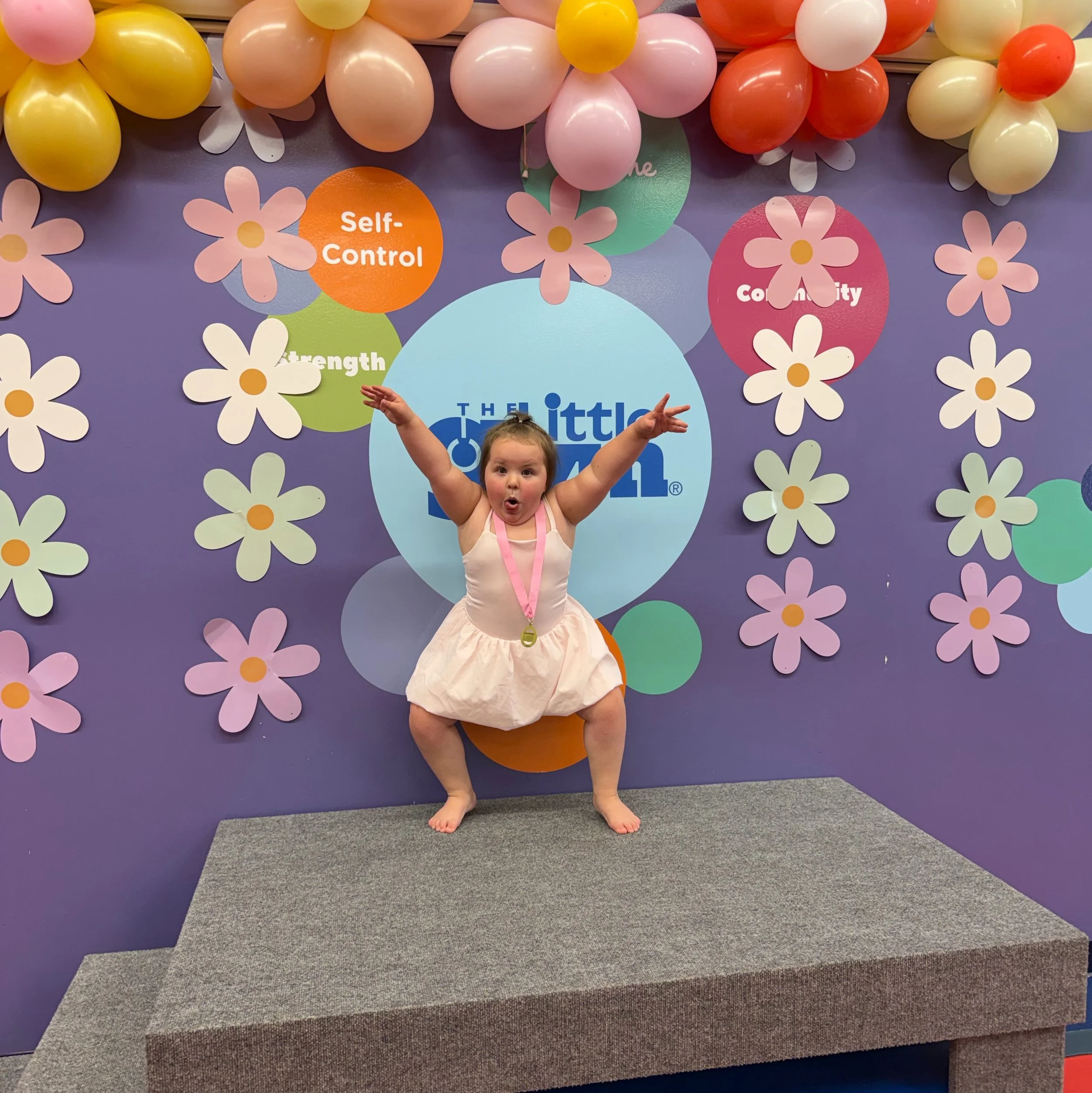 Bella Grace striking a playful pose on stage in her gymnastics leotard at The Little Gym, capturing her joyful personality, confidence, and love for making people smile.