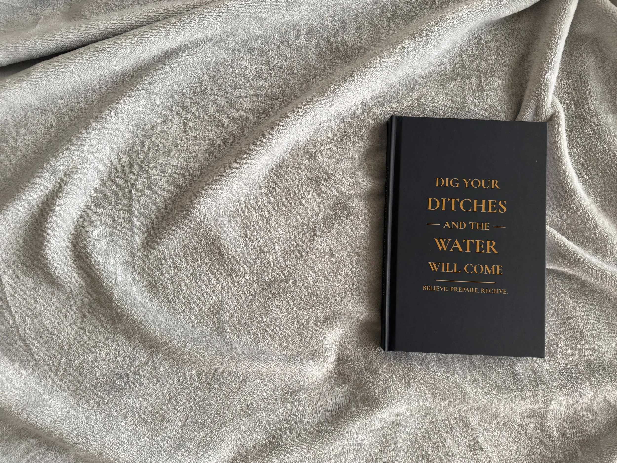 A black book with gold text that reads 'Dig Your Ditches and the Water Will Come' lies on a cream-colored, textured fabric surface.