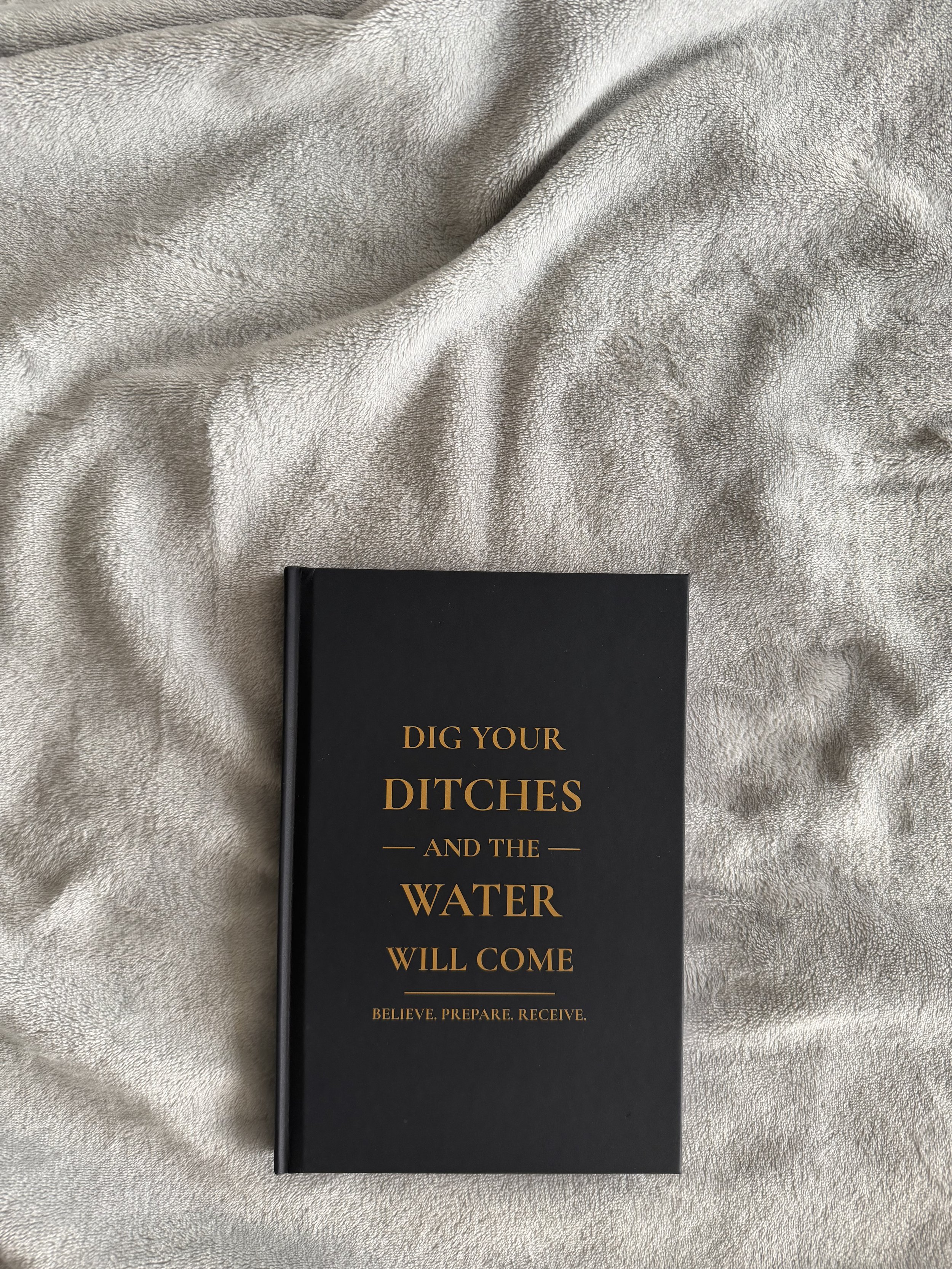 A black book with gold lettering that reads "Dig Your Ditches and the Water Will Come" placed on a light-colored fabric background.