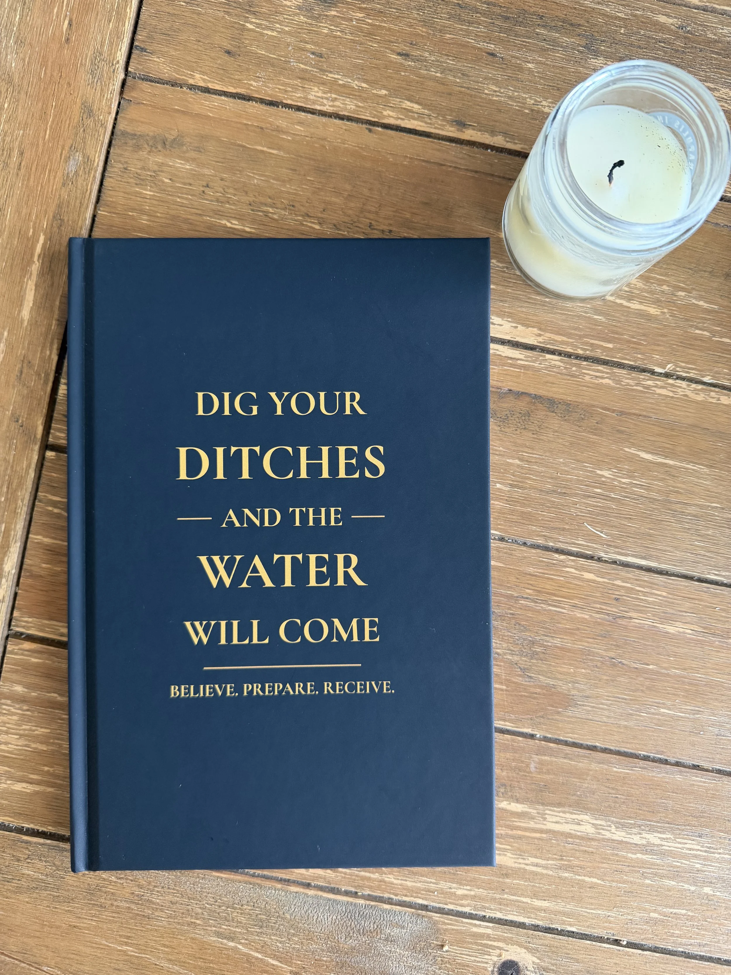 A blue book with the title "Dig Your Ditches and the Water Will Come" on a wooden table, next to a lit white candle in a glass jar.