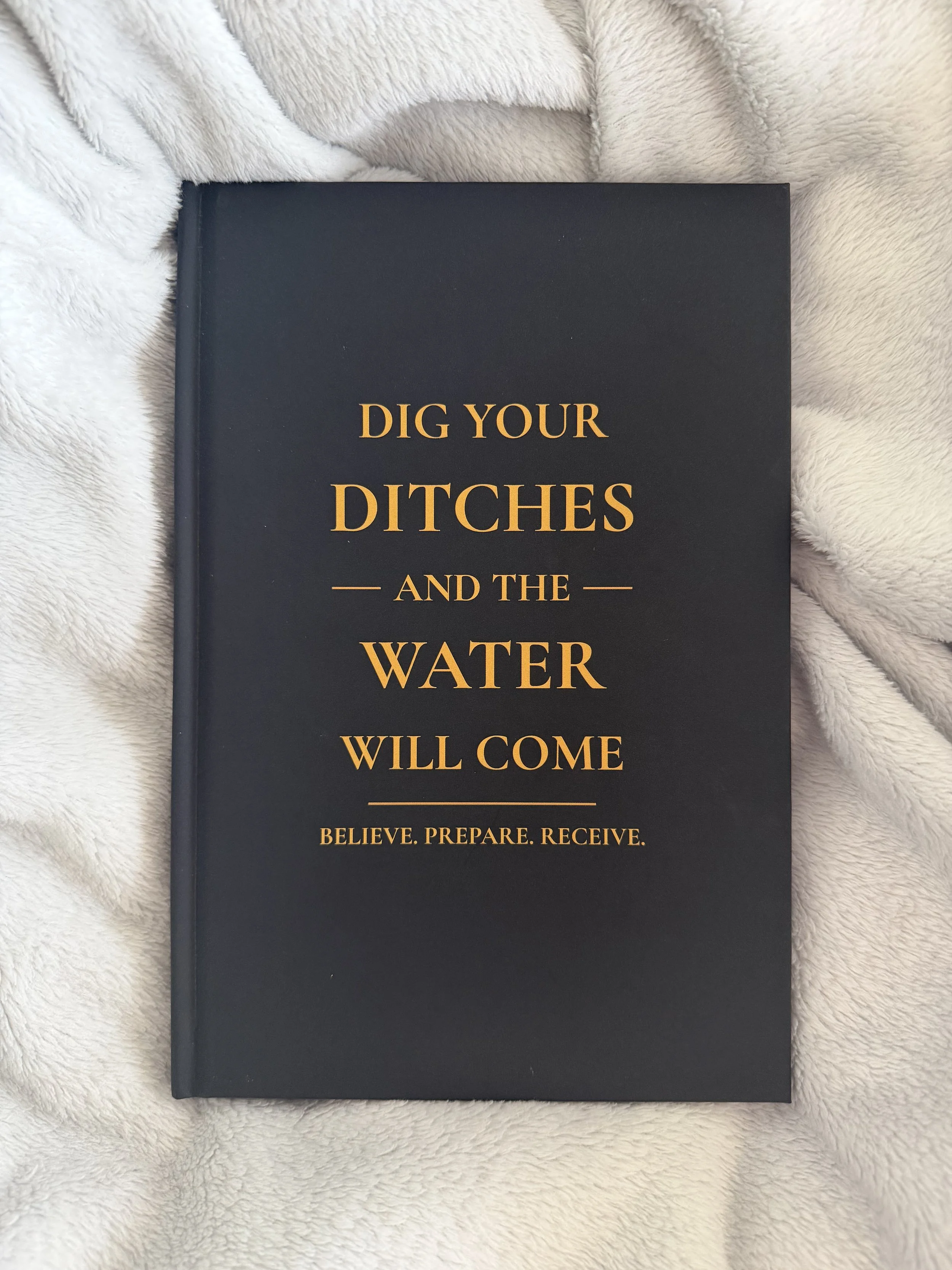A hardcover book titled "Dig Your Ditches and the Water Will Come" with the subtitle "Believe. Prepare. Receive." on a soft, beige fabric background.