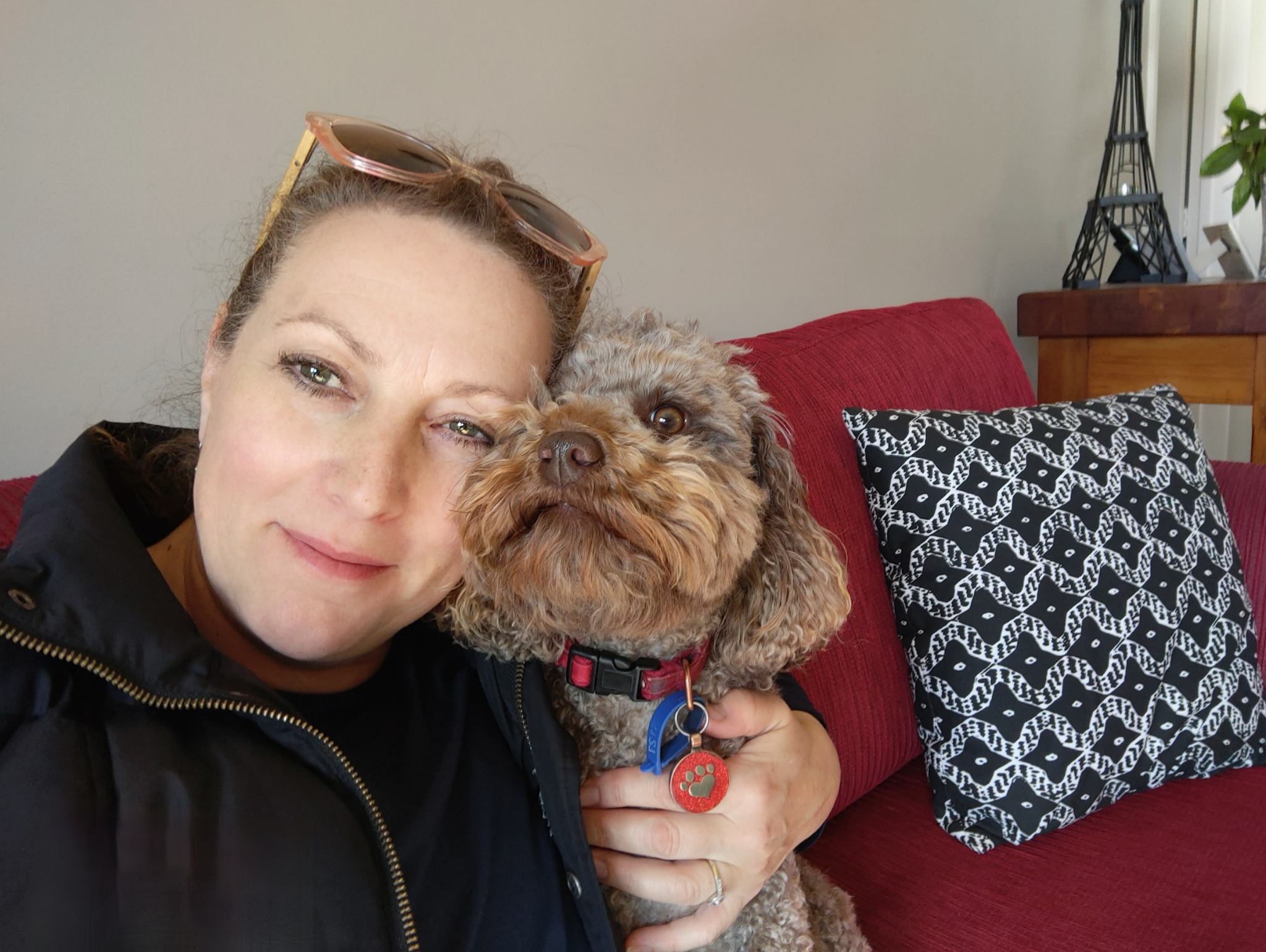 A woman with sunglasses on her head and a black jacket, smiling, holding a small curly-haired dog with brown and tan fur, sitting on a red couch with a patterned black and white pillow, in a room with a beige wall, a wooden side table, and a decorative metal Eiffel Tower.
