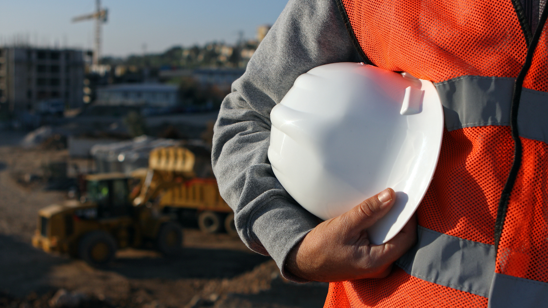 Construction worker holding a white helmet at a construction site with machinery and buildings in the background.