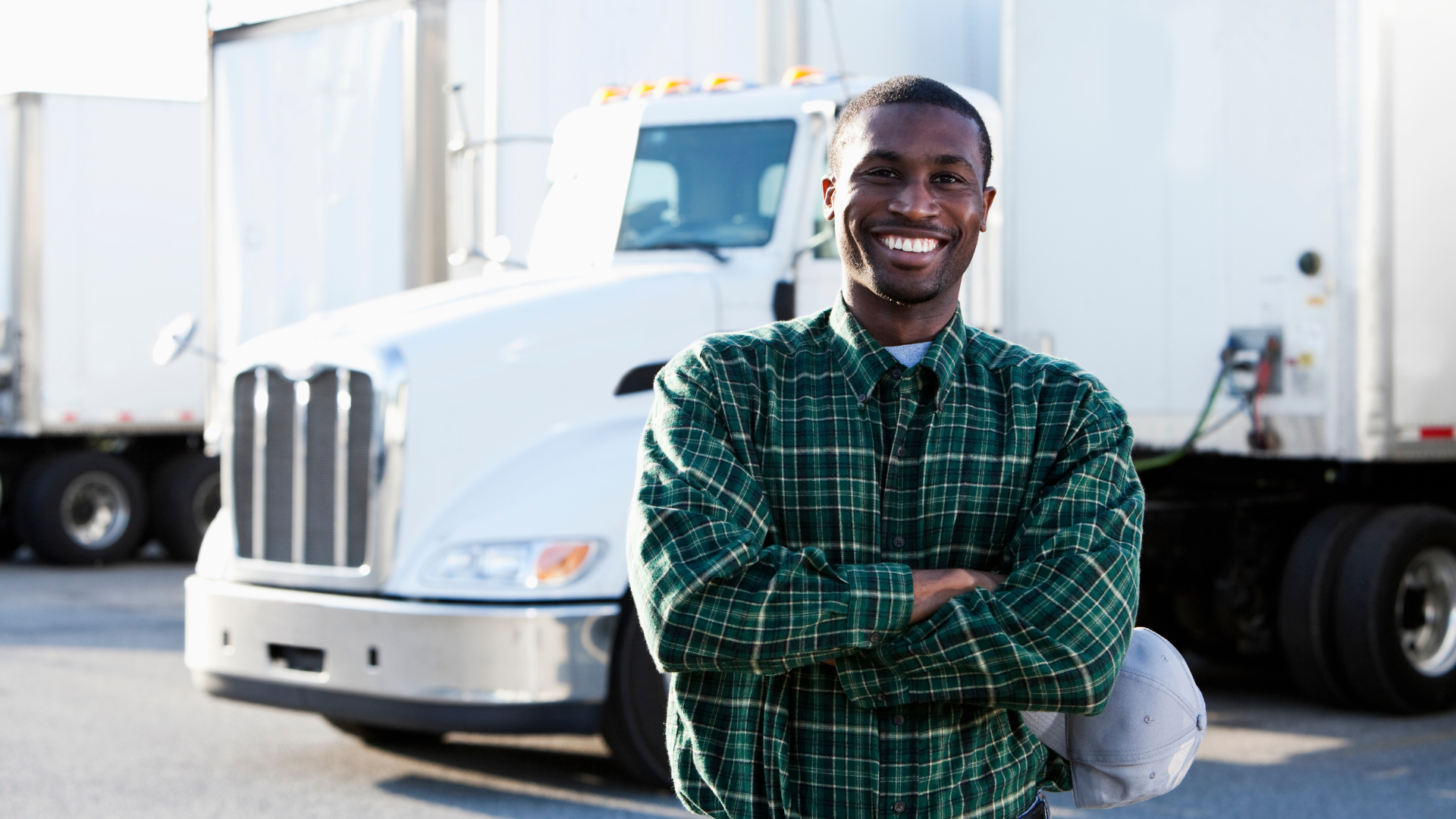 A smiling man in a green plaid shirt standing with crossed arms in front of a white semi-truck, holding a white helmet with a face shield.