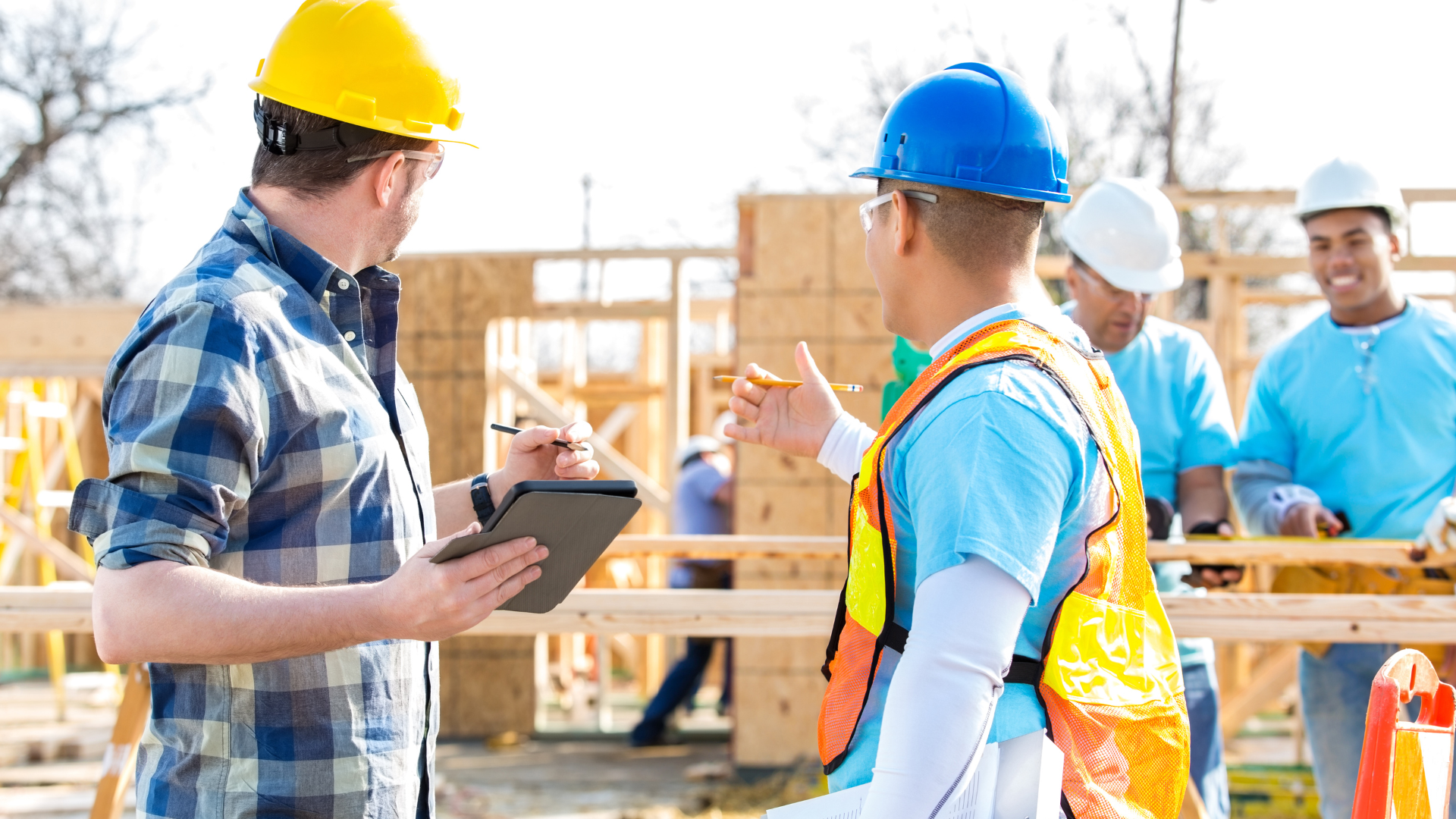 Construction site with workers wearing hard hats. One worker in a yellow hat discusses with another in a blue hat and safety vest. Others work in the background.