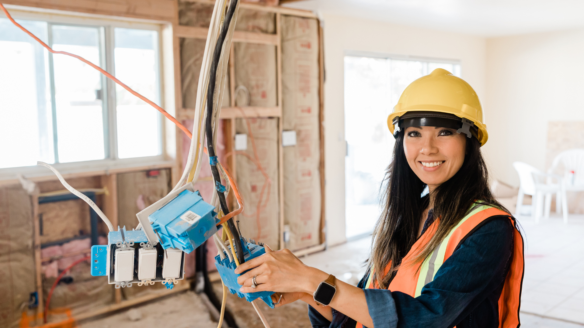 A woman wearing a yellow hard hat and safety vest working on electrical wiring at a construction site inside a house.