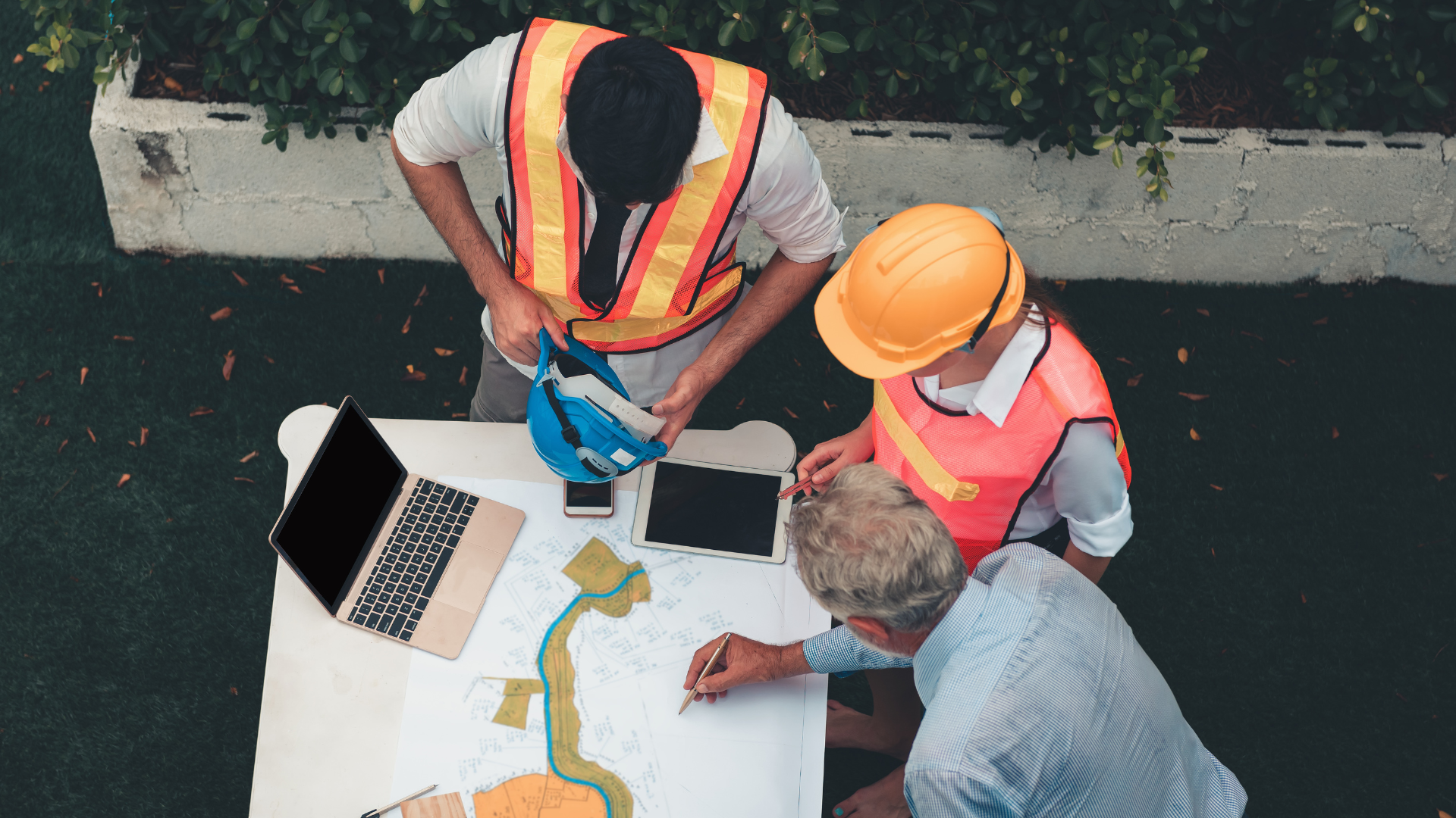 A group of three people, two men wearing construction helmets, are gathered around a table outdoors. The table has a map, a laptop, a tablet, a smartphone, and some papers. One man is holding a blue helmet and a pen, and they appear to be discussing a project or plan.