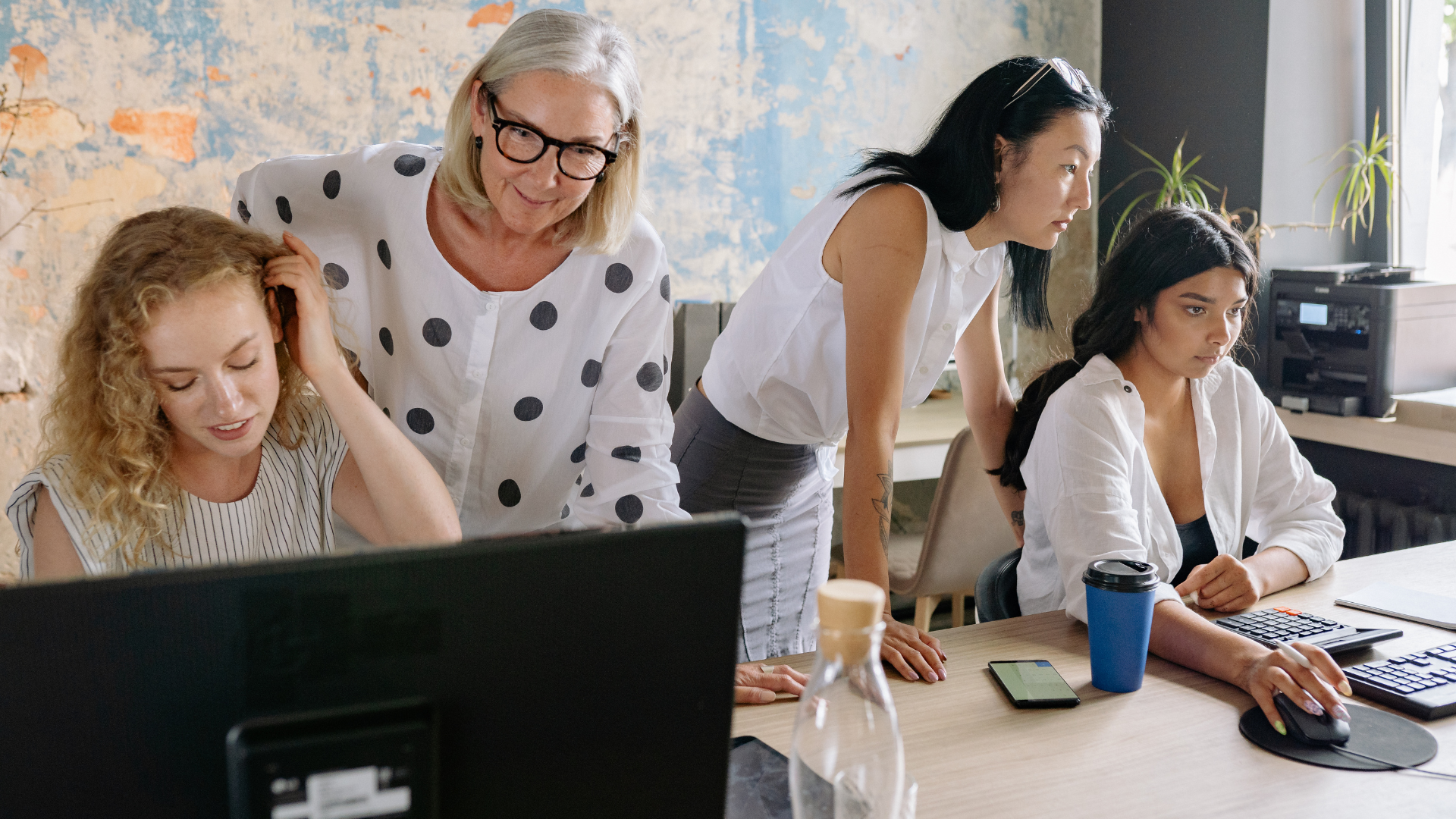 Group of four women working together in an office, looking at a computer screen, some using keyboards, in a room with a brick wall and large window with potted plants.