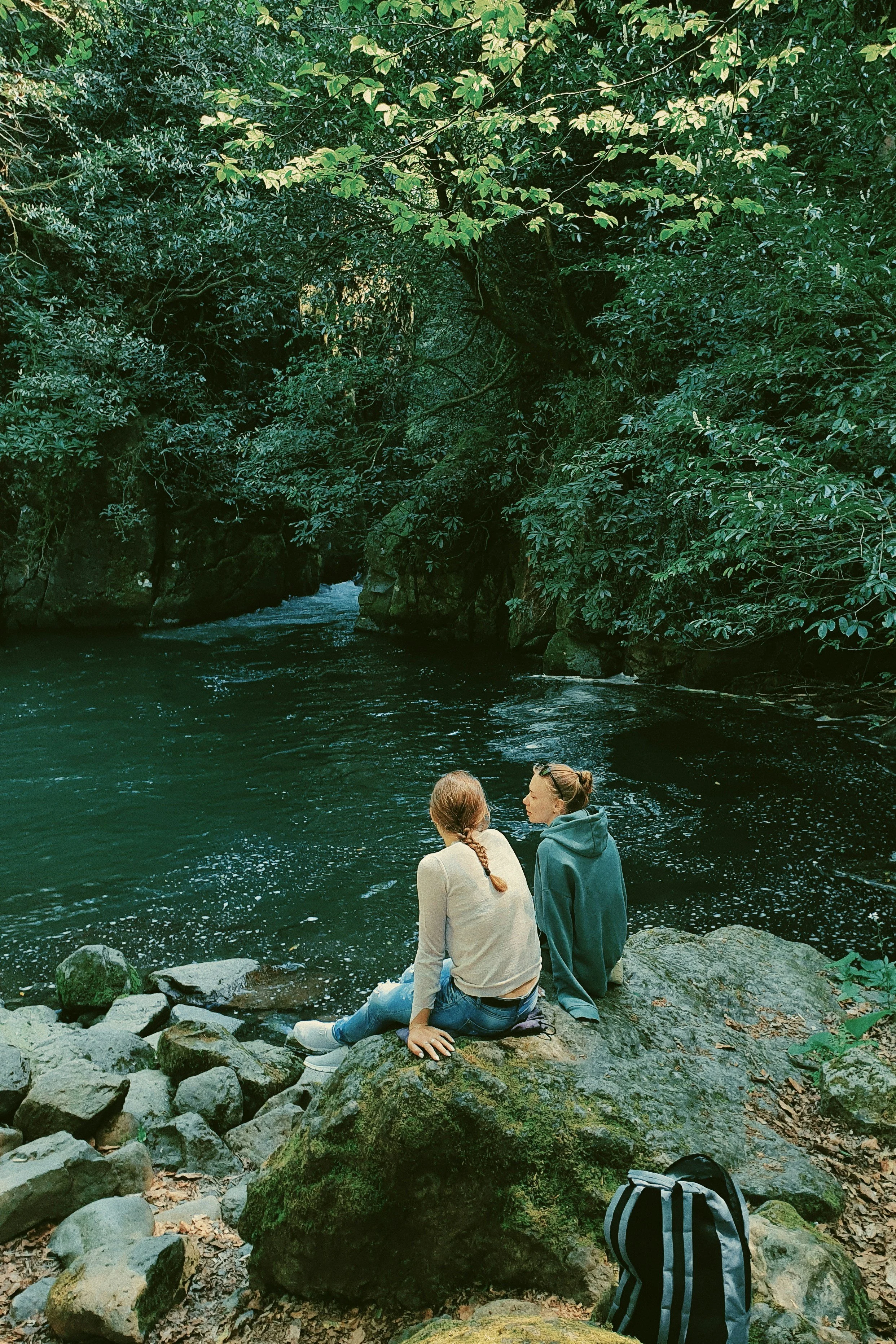 Two women sitting on rocks beside a river in a forested area, with lush green trees overhead.