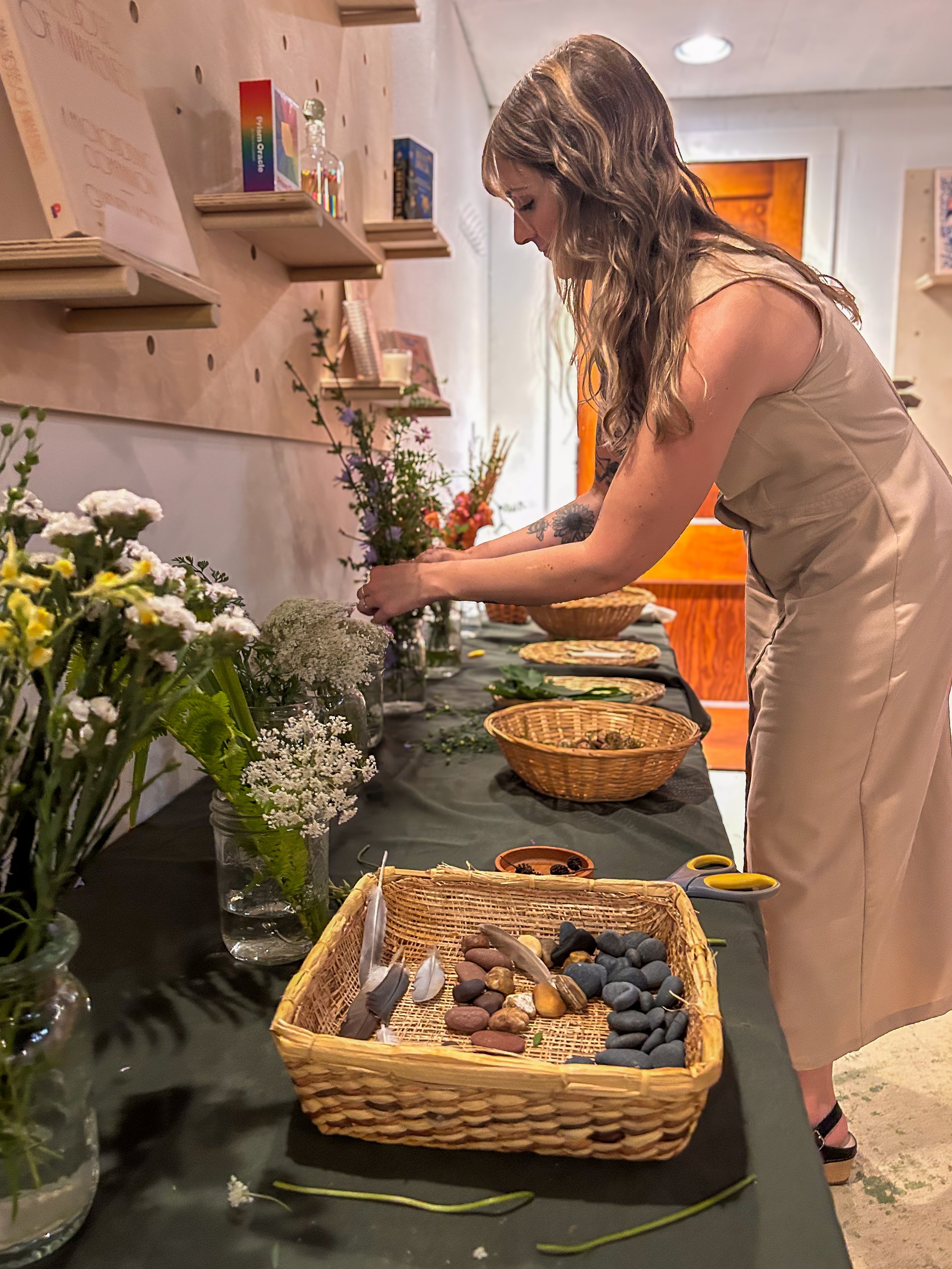 Halie Tenor, founder of nested kin, arranging flowers at a table with various vases of flowers, stones, and decorative items. Shelves with books and decorative objects are on the wall behind her.