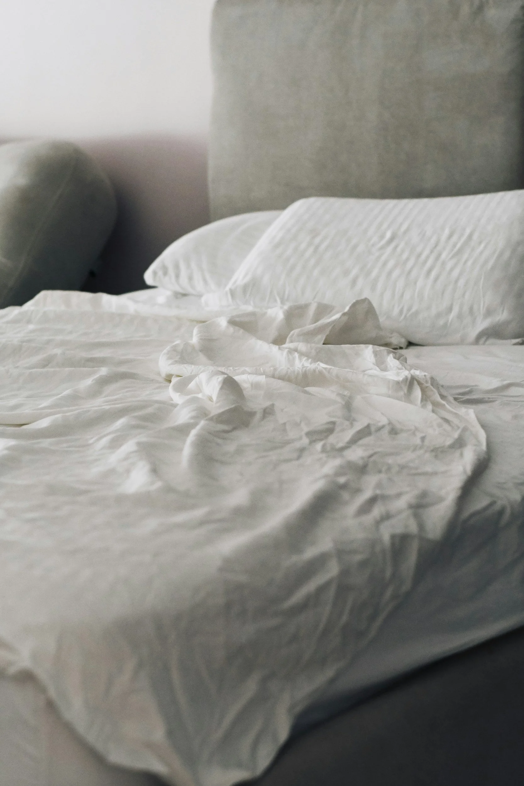 A crumpled white bedsheet with matching white pillows on a bed, with a light-colored headboard and a pillow leaning against it.