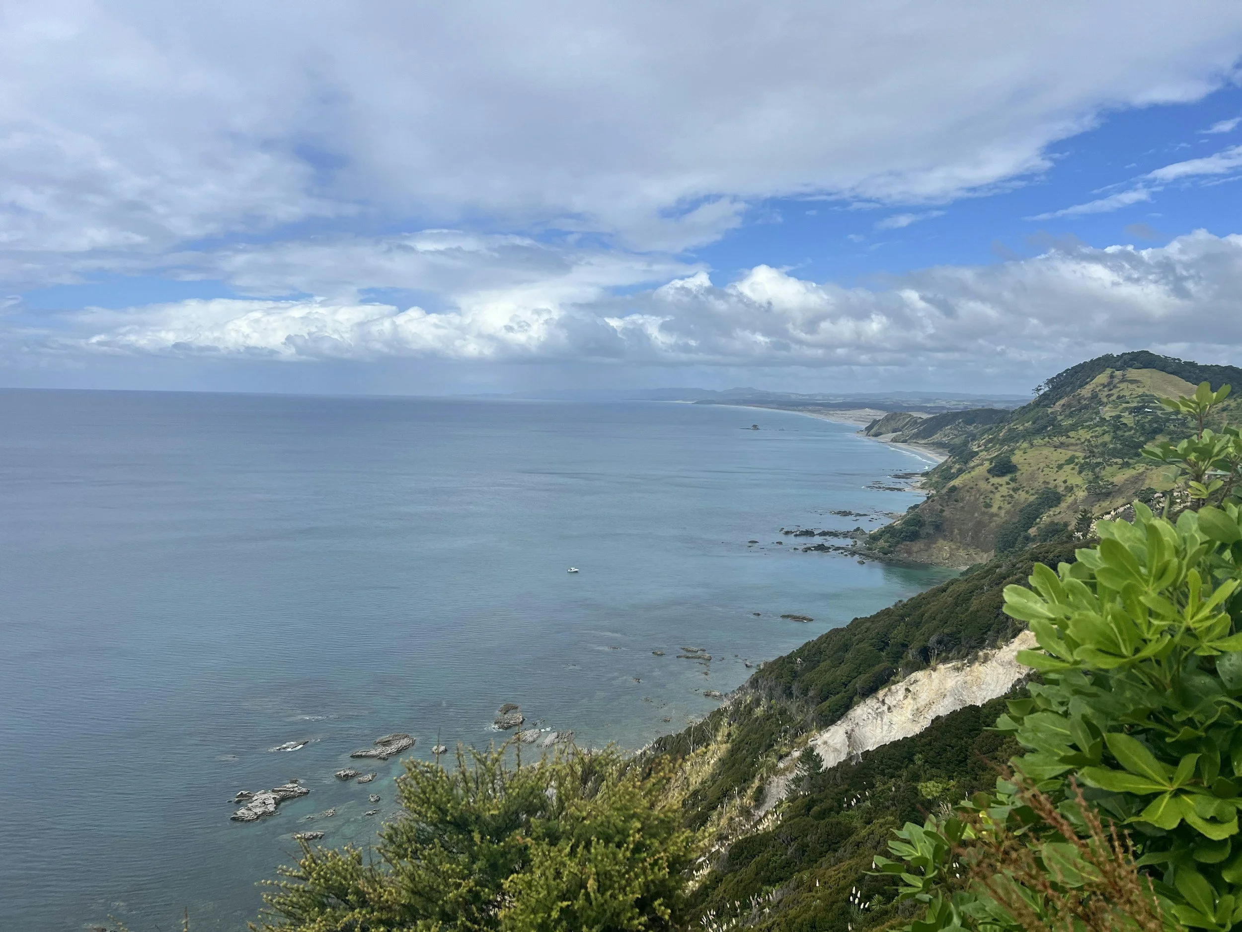 Scenic view of a coastline with a green hill on the right and calm ocean water on the left under a partly cloudy sky.