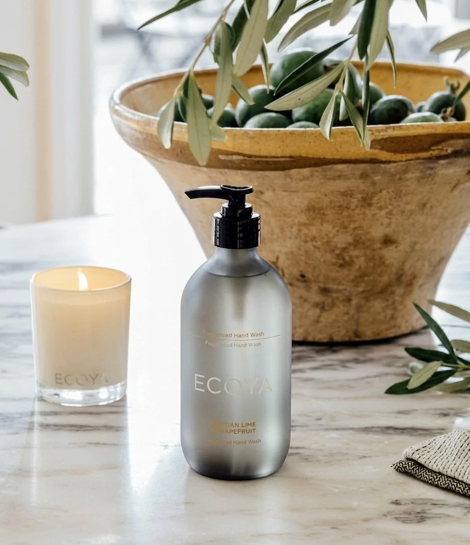 A frosted bottle of ECOYA hand wash with a pump dispenser, a lit candle, a large ceramic bowl with green olives and olive branches, on a marble countertop.