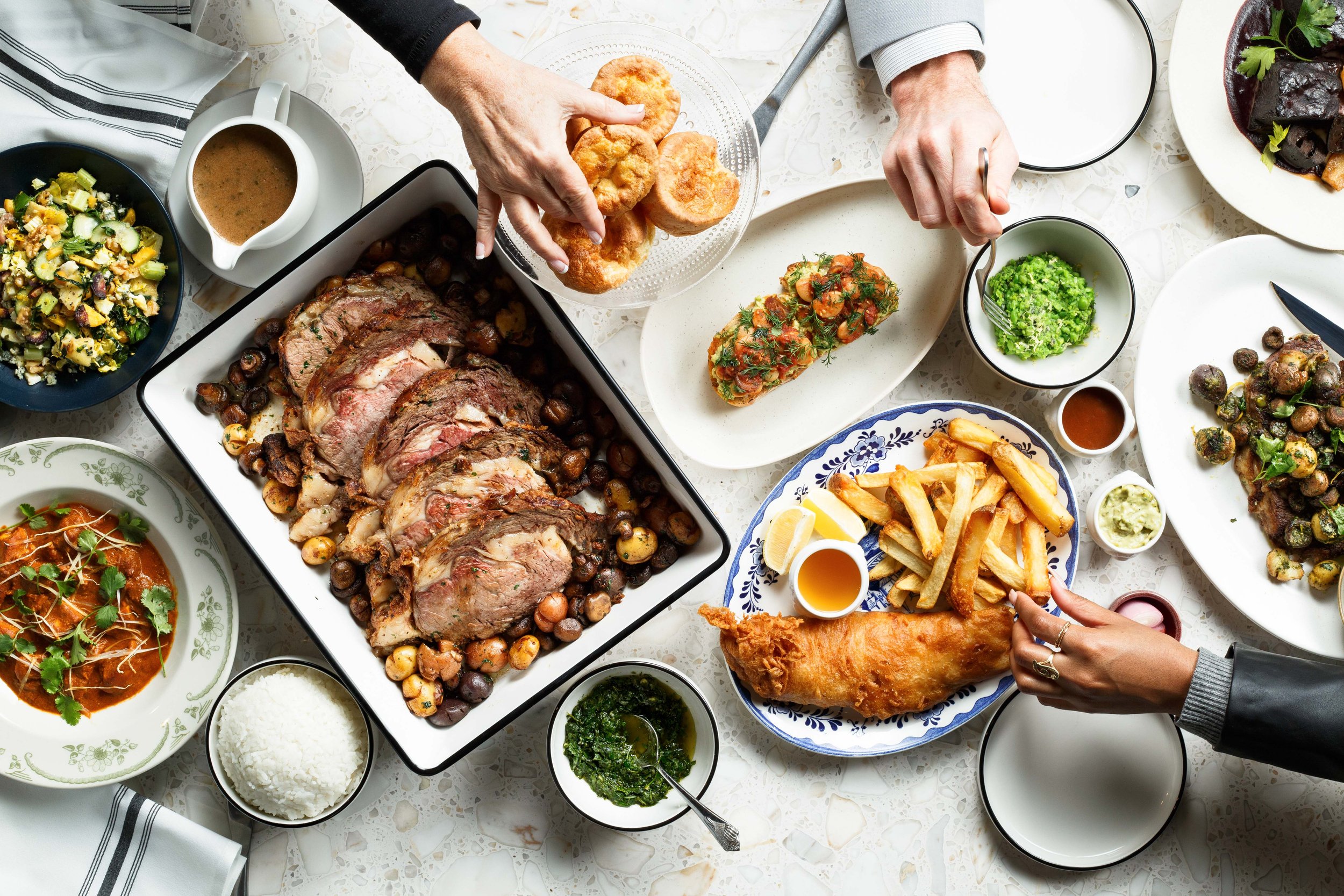 Commercial lifestyle photo of a full English brunch spread with hands reaching over the table, shot for a hospitality brand campaign.