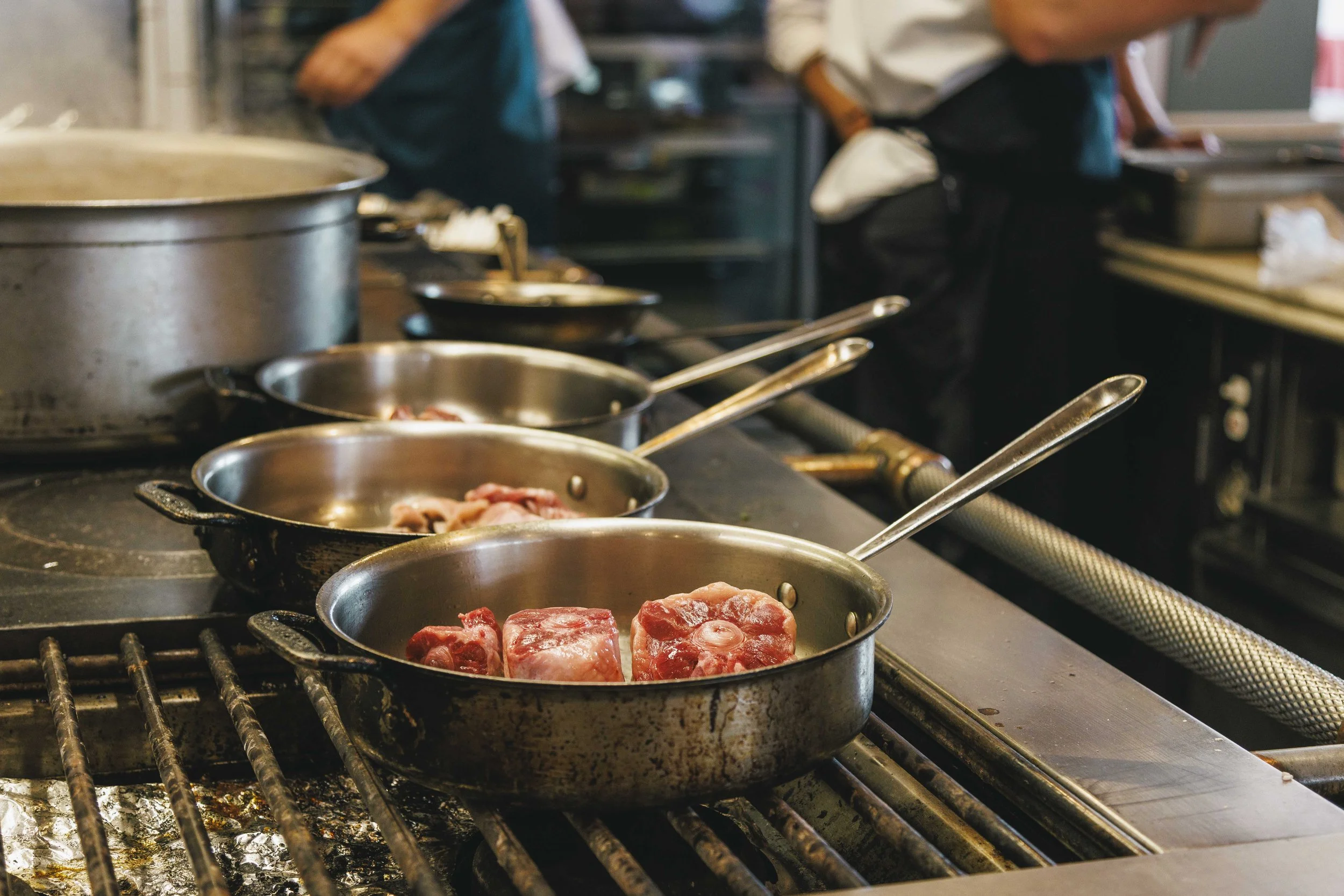 Multiple stainless steel pans with raw meat cooking on a commercial stove in a busy kitchen with two chefs in the background.