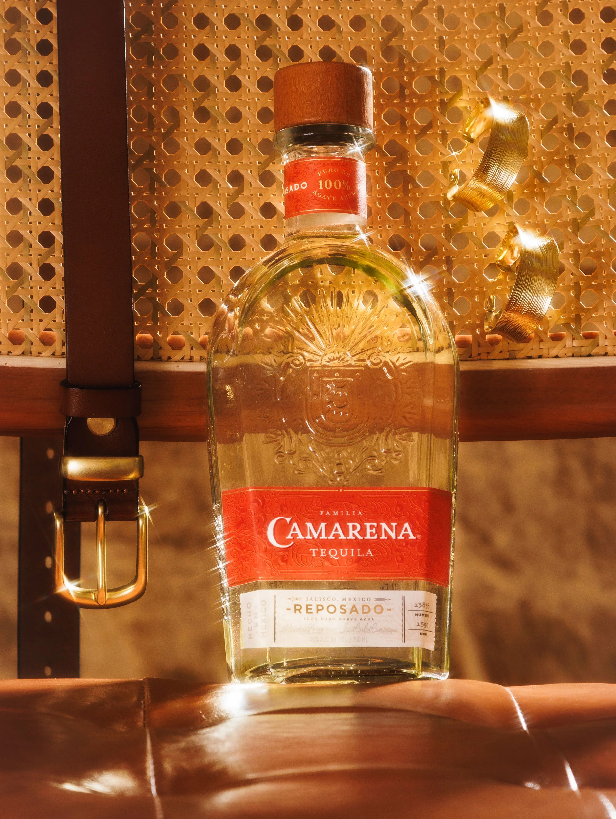 A glass bottle of Camarena Reposado tequila with a wooden cap, placed on a wooden surface against a decorative woven background, with golden earrings hanging to the right and sunlight reflecting off the bottle.