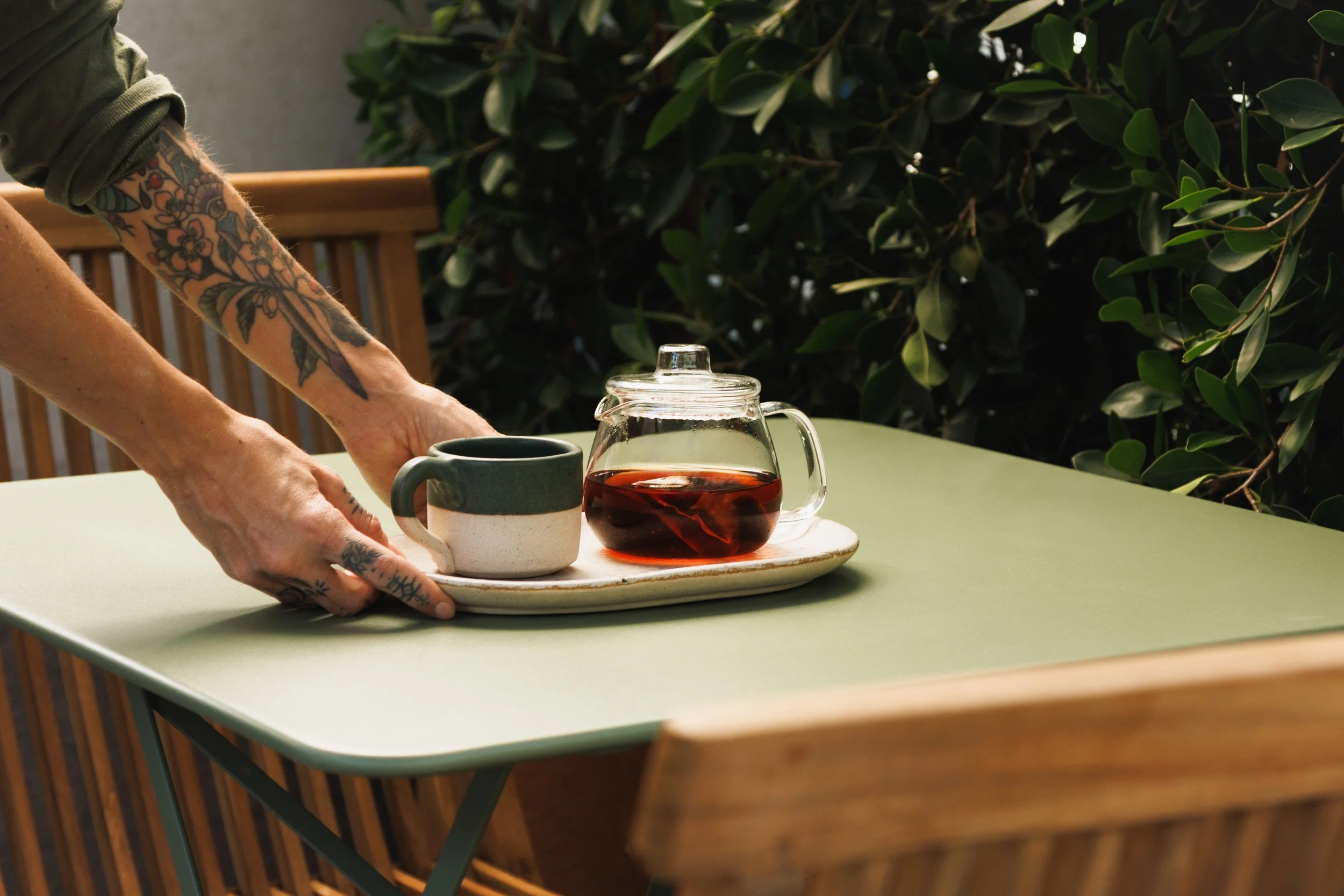 Atmospheric environmental lifestyle photo of a waitress placing a pot of tea on a table with vegetation around, for a hospitality campaign.
