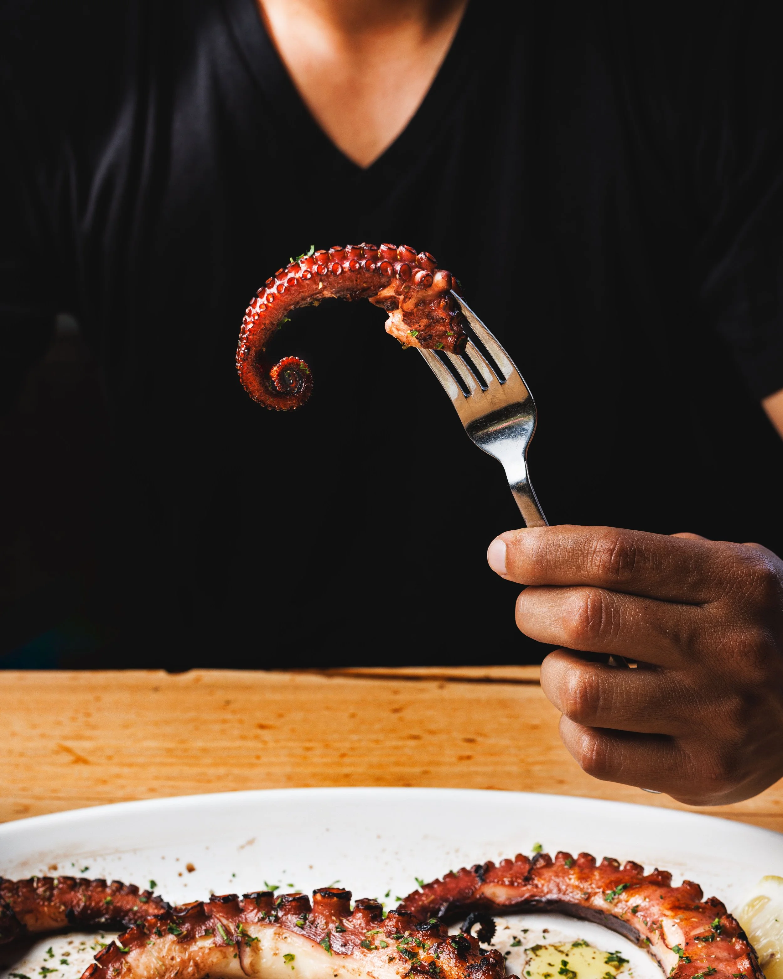 Commercial photo of a person wearing a black shirt holding a fork with a piece of grilled octopus tentacle over a white plate with more octopus and garnishments.