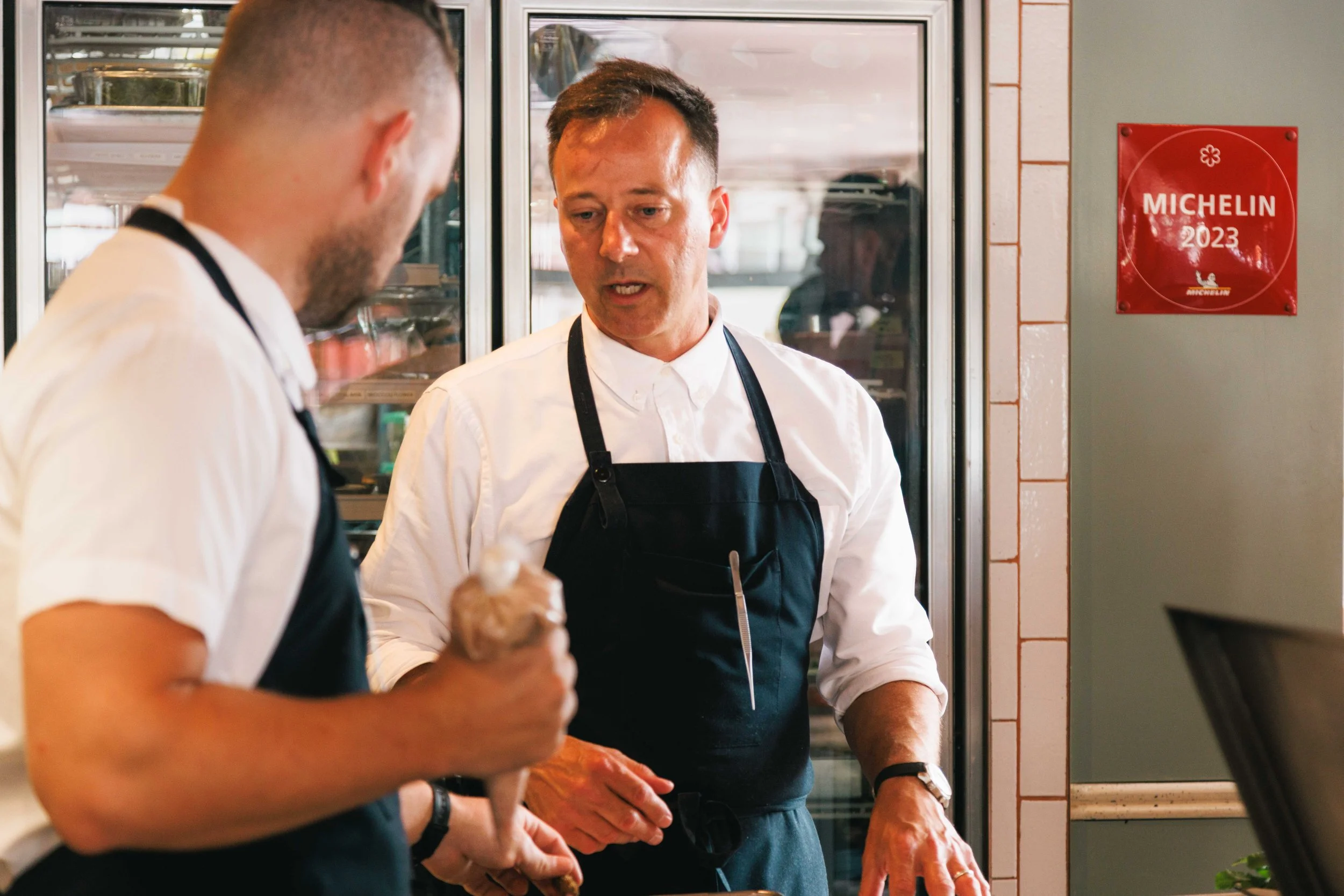 Editorial hospitality photo of two chefs speaking in the kitchen with a Michelin award on the wall.