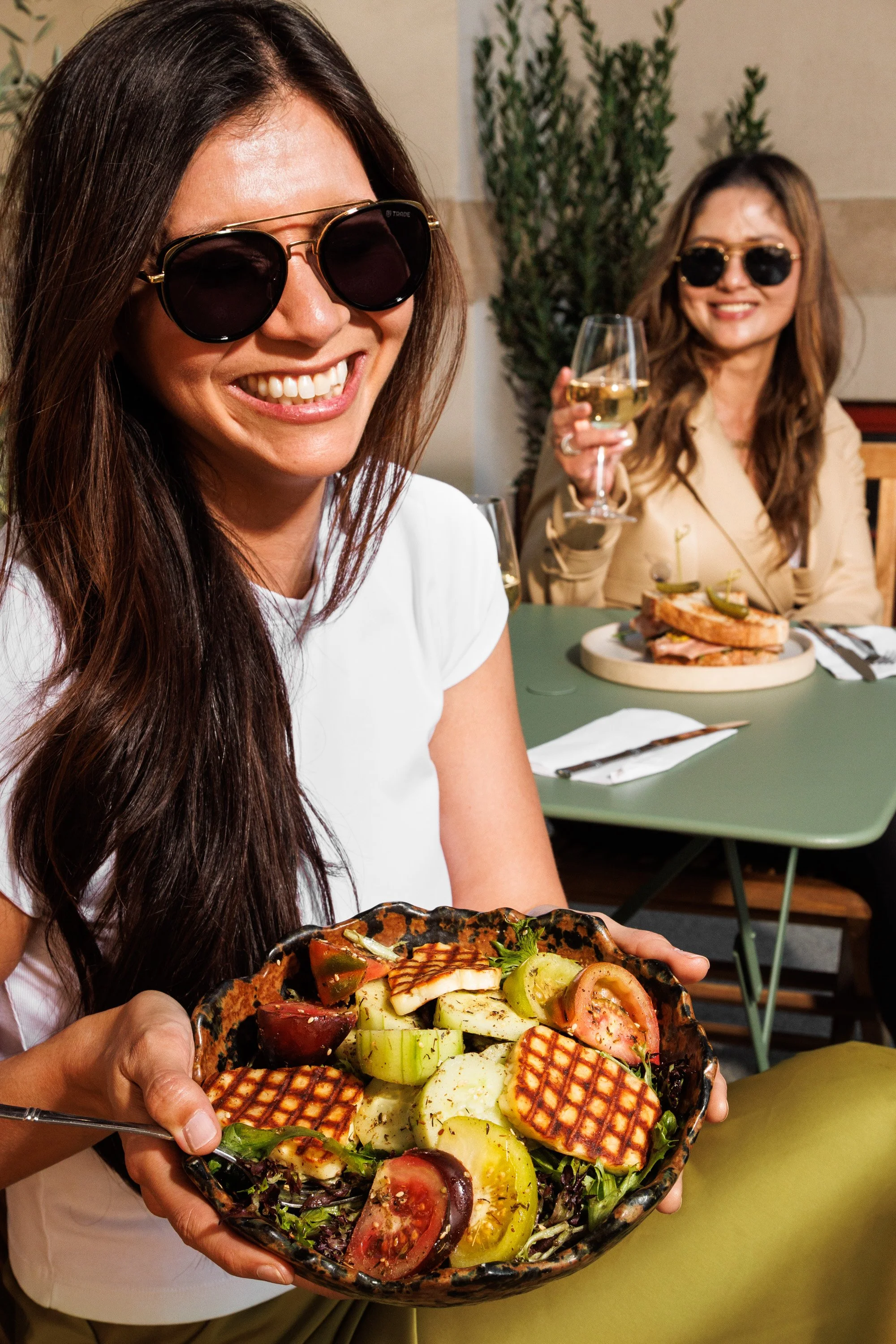 Two women smiling in a restaurant, one holding a vegetable and grilled cheese salad in a bowl, the other holding a glass of white wine, with a plate of sandwich on the table.