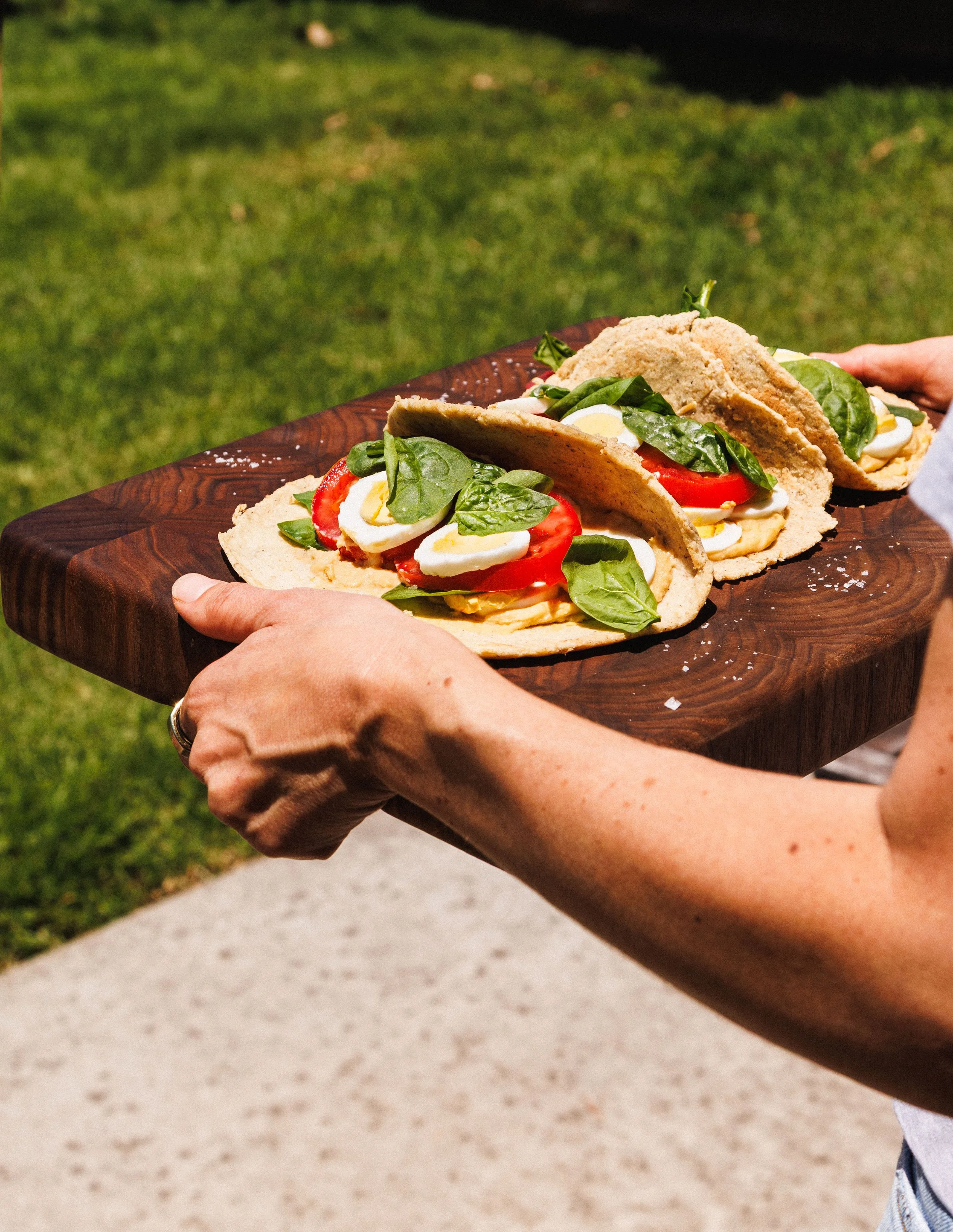 Commercial photo of a person holding a wooden platter with tacos, featuring sliced tomatoes, scrambled eggs, spinach, and other fillings, outdoors on a grassy area.