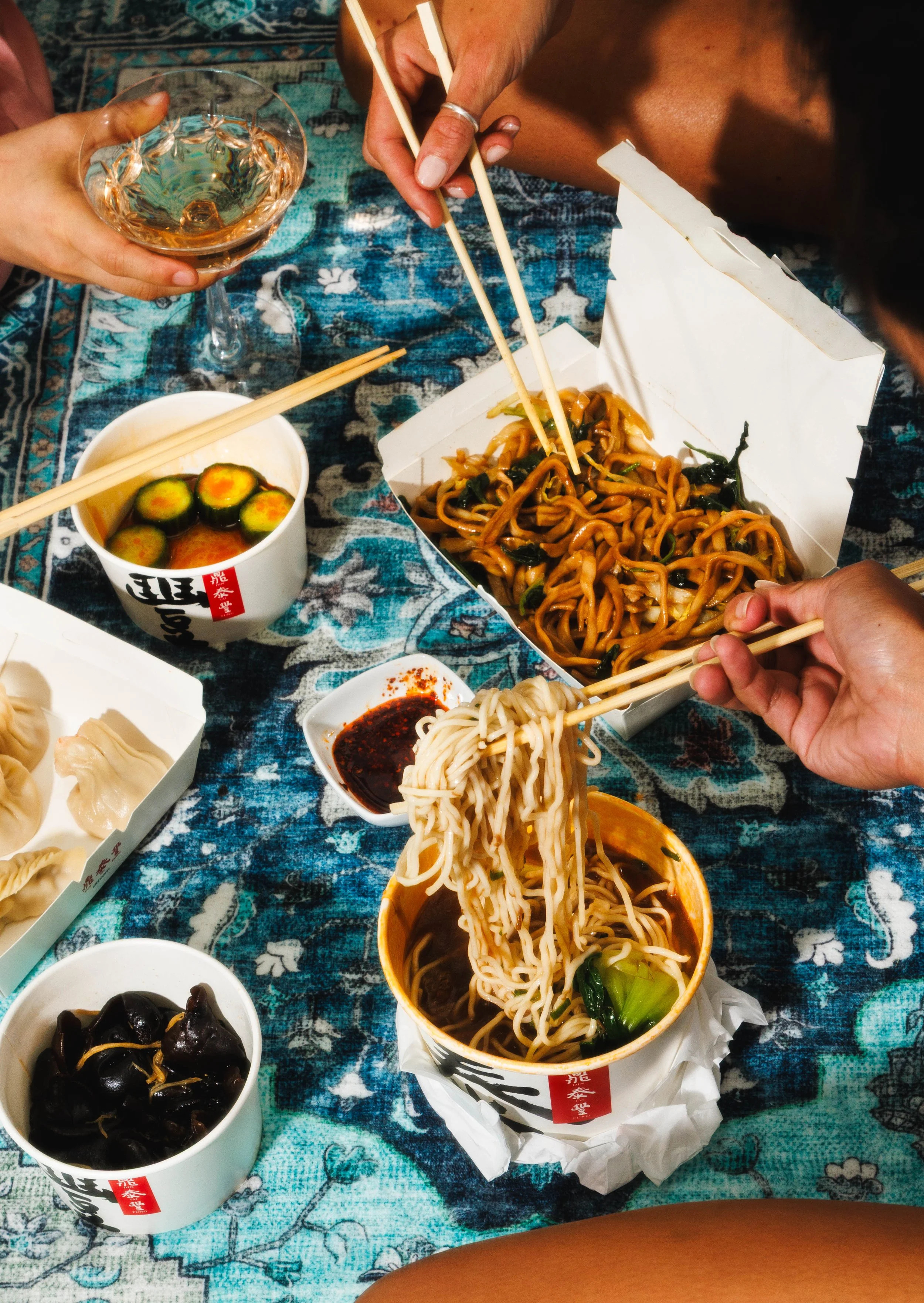 Commercial food photo of girls eating Chinese noodles on a blue patterned rug for a campaign.