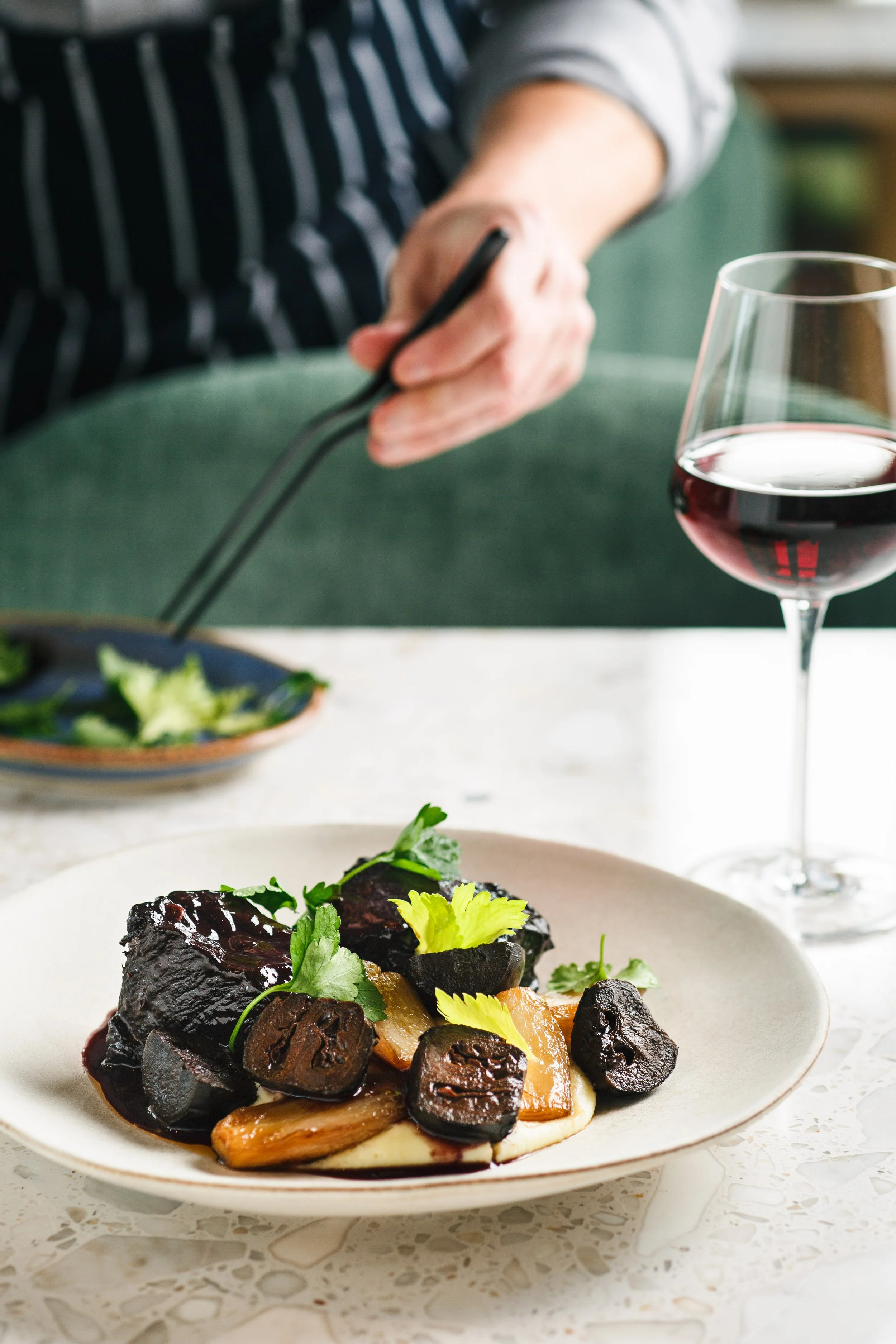 Commercial food photo of a chef plating a braised beef dish and red wine for a hospitality brand campaign.