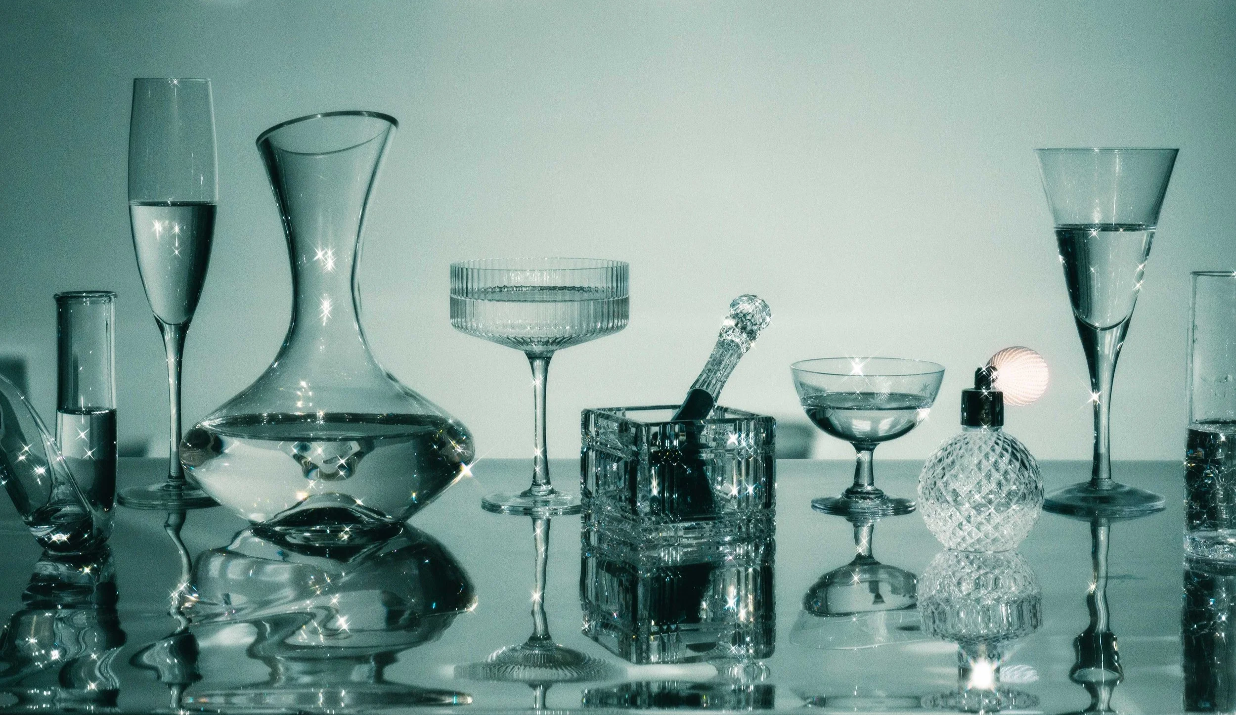 Commercial still life photo of glassware arranged on a reflective surface with light reflections and sparkles.