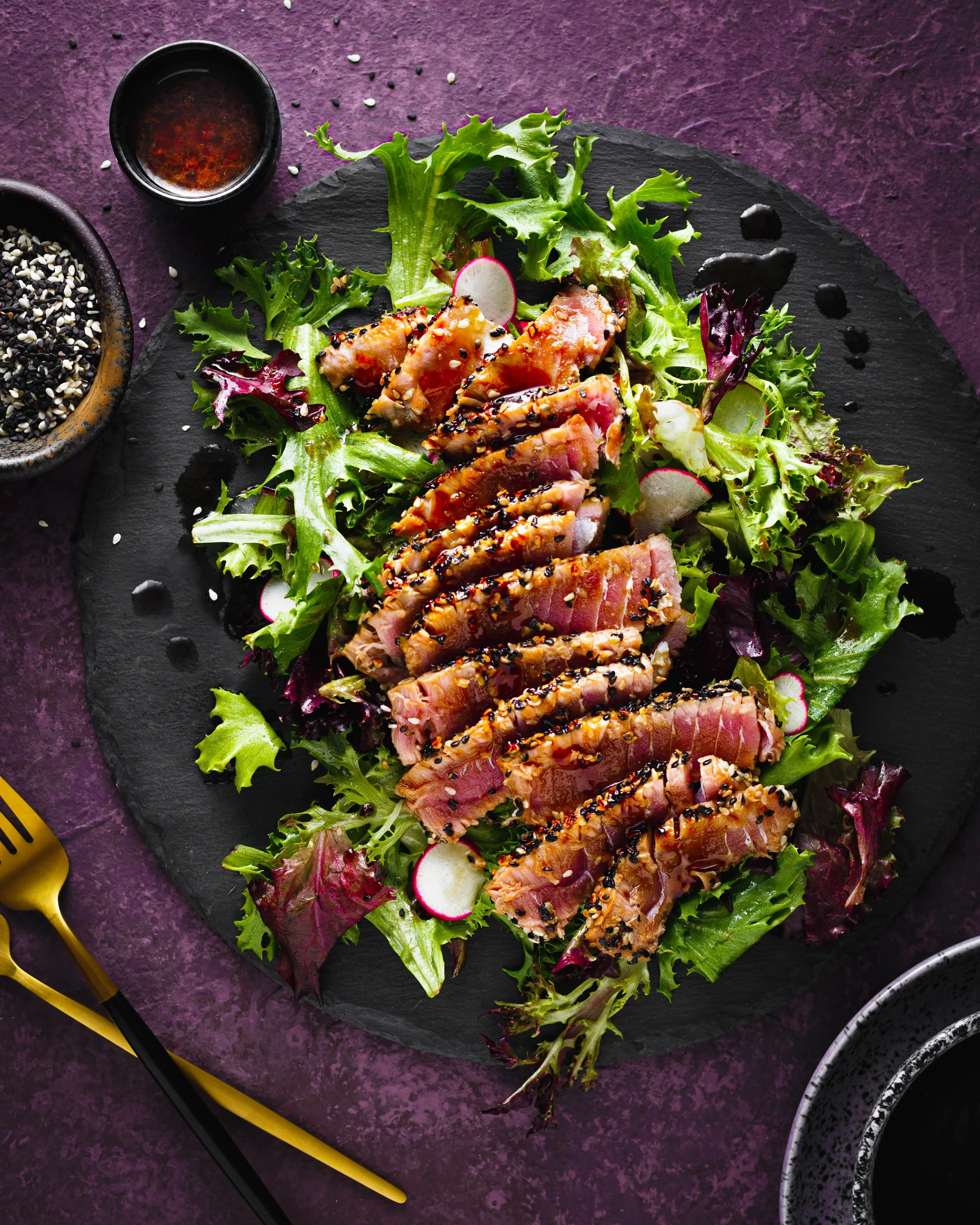 Commercial food photo of sliced raw tuna topped with black and white sesame seeds on a bed of mixed greens, radish slices, and garnishes, served on a black slate plate with soy sauce and bowls of sesame seeds around it.