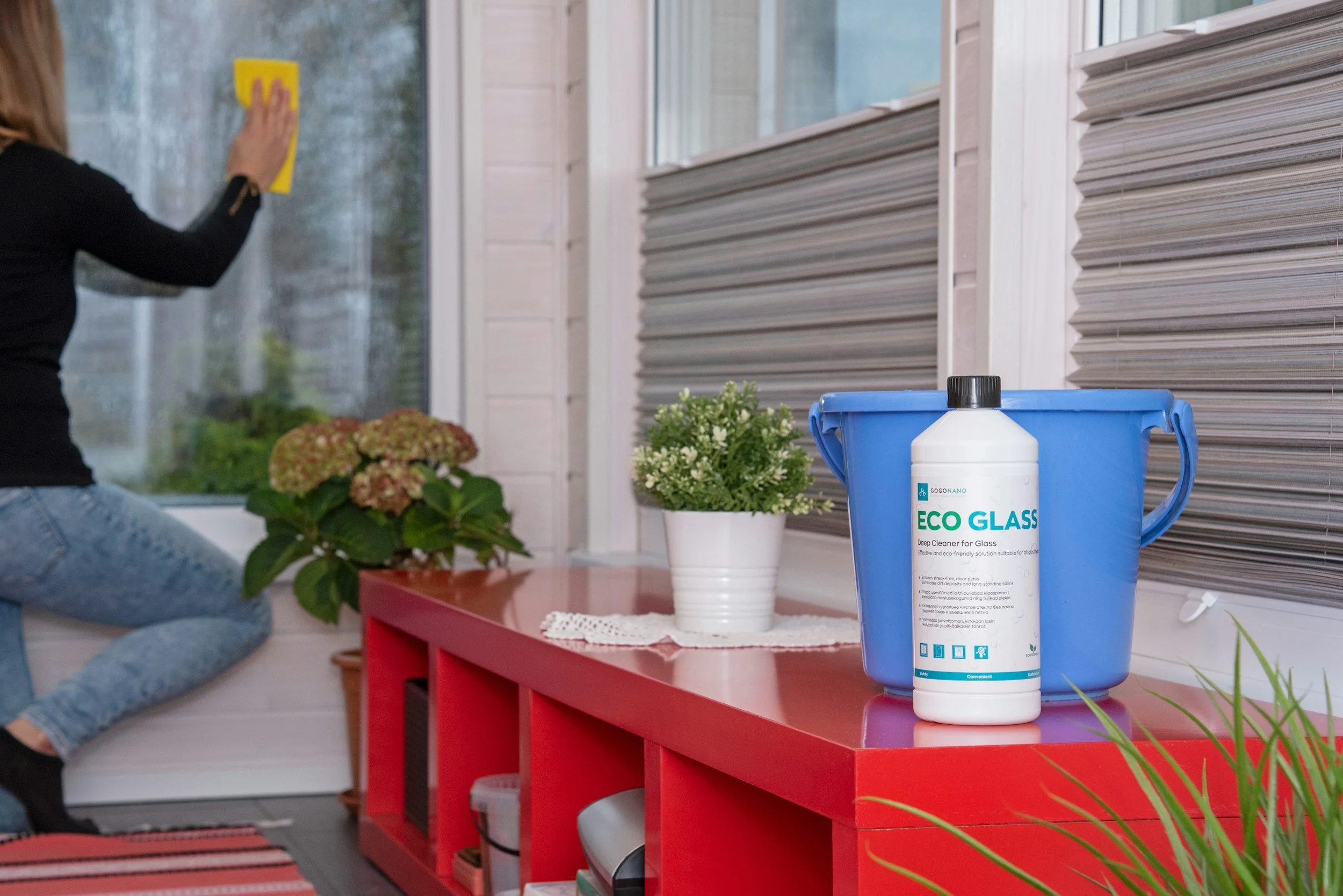 A person is cleaning a window with a yellow cloth, seated beside a red table with potted plants, a blue container labeled "Eco Glass," and a window with brown blinds.