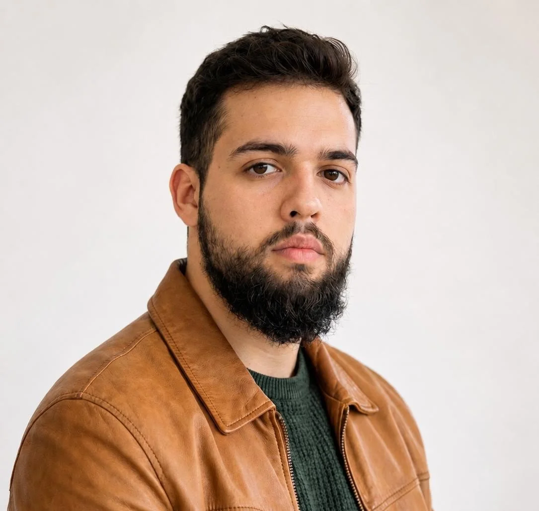 Homem de pele clara, cabelo castanho escuro e barba, vestindo jaqueta de couro marrom e suéter verde, contra fundo branco.
