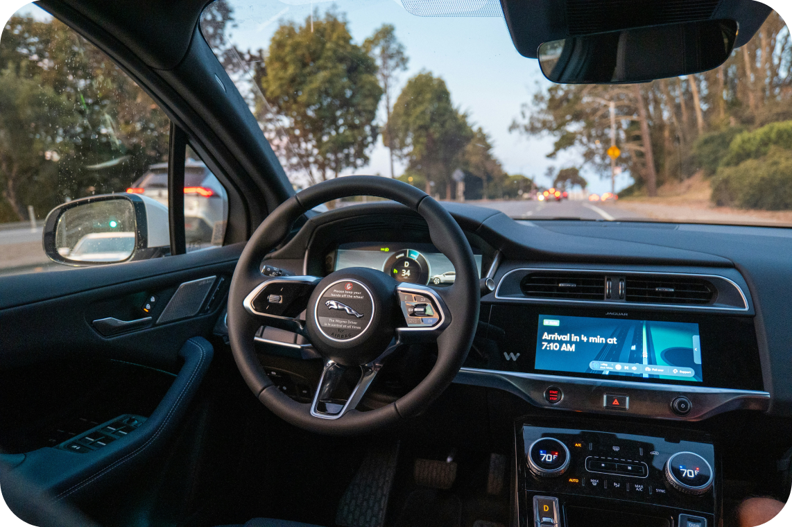 The interior of a modern Jaguar electric vehicle, showing the steering wheel with the Jaguar logo, digital instrument cluster, infotainment screen displaying a navigation alert, and climate controls, with a view of a road and trees through the windshield.